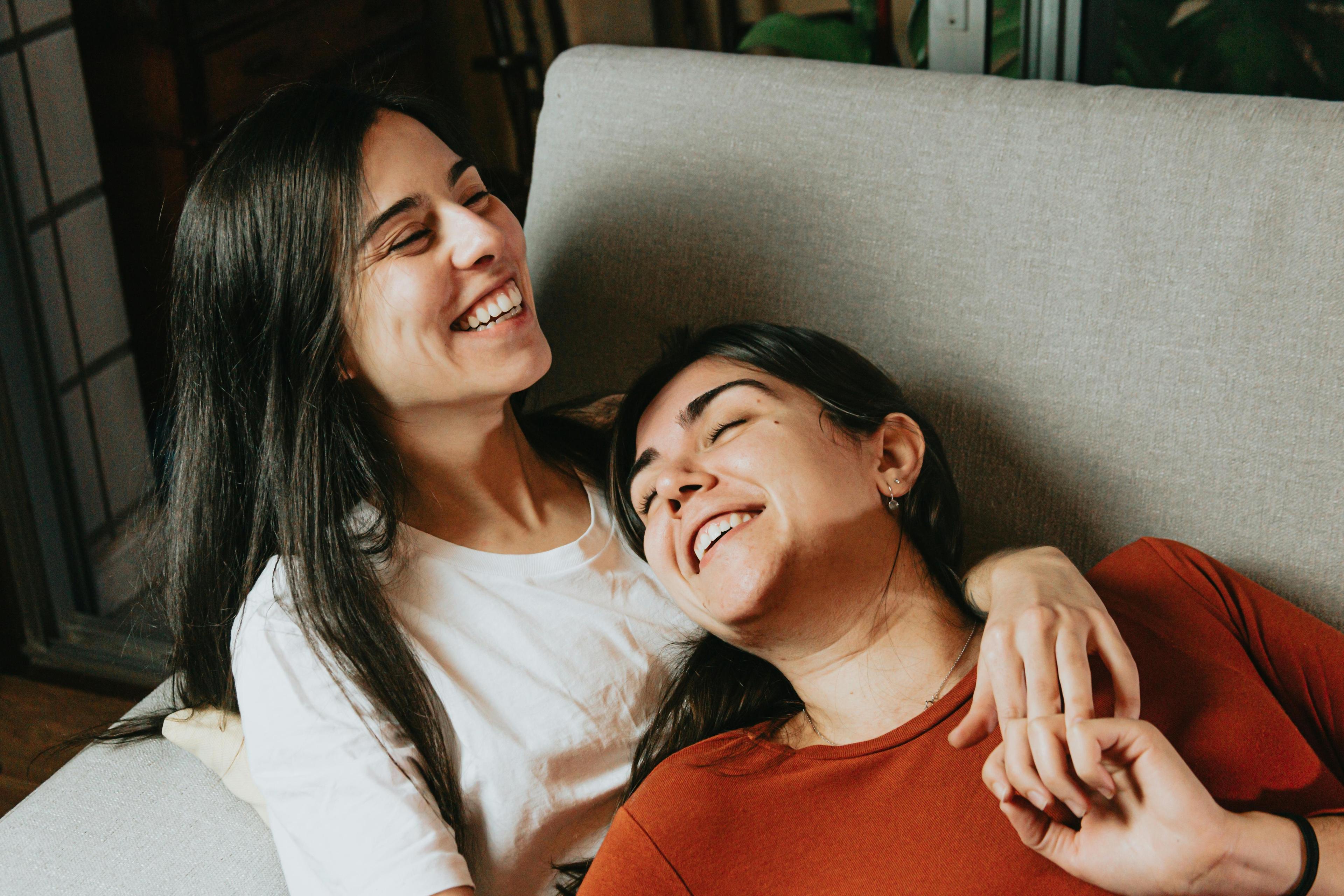 Two women laughing abut different stages of relationships, lying on a couch together, sharing a moment of comfort and companionship