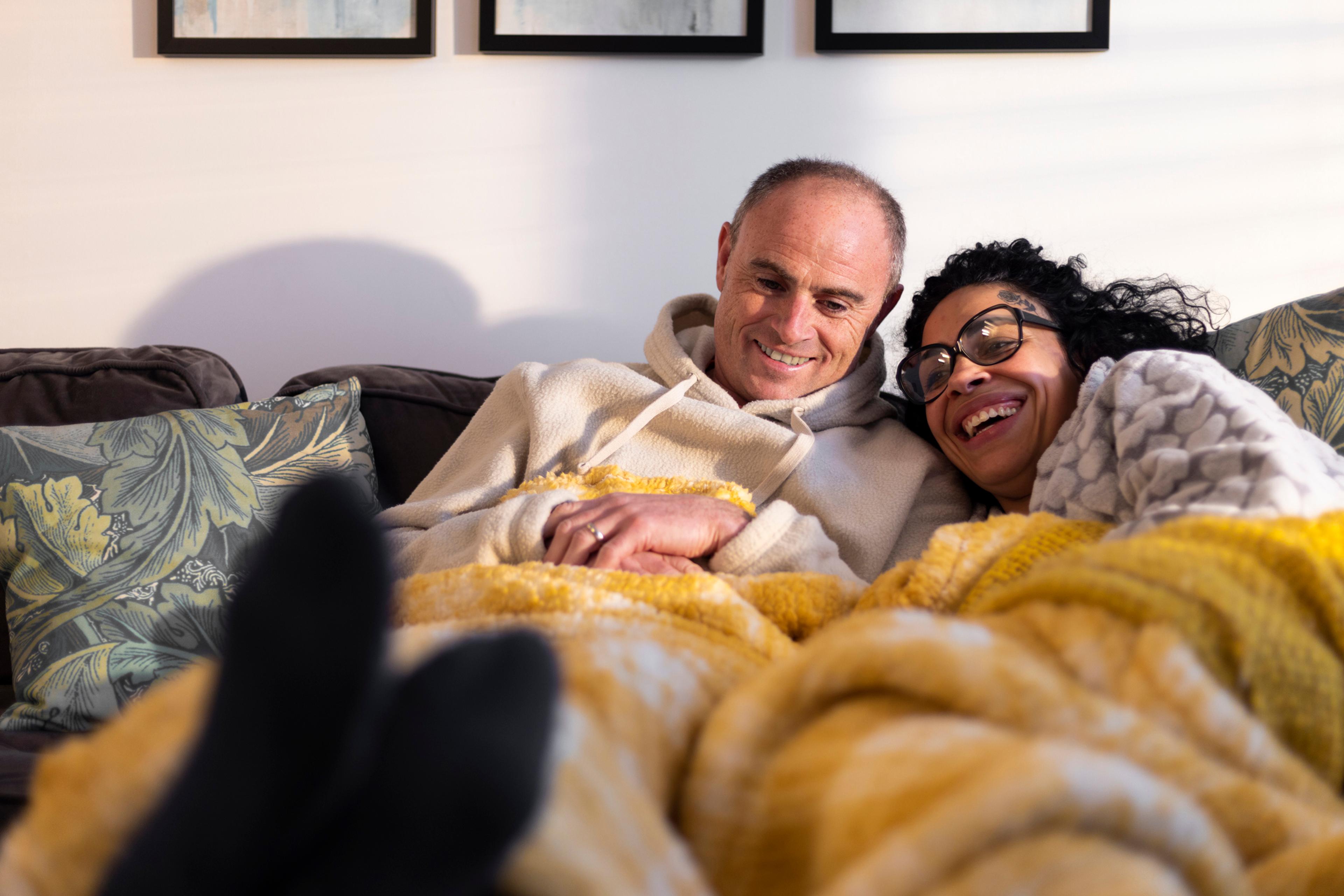 A woman and a man sitting in a couch with a yellow blanket.  They laugh as they do the magical five hour exercise.