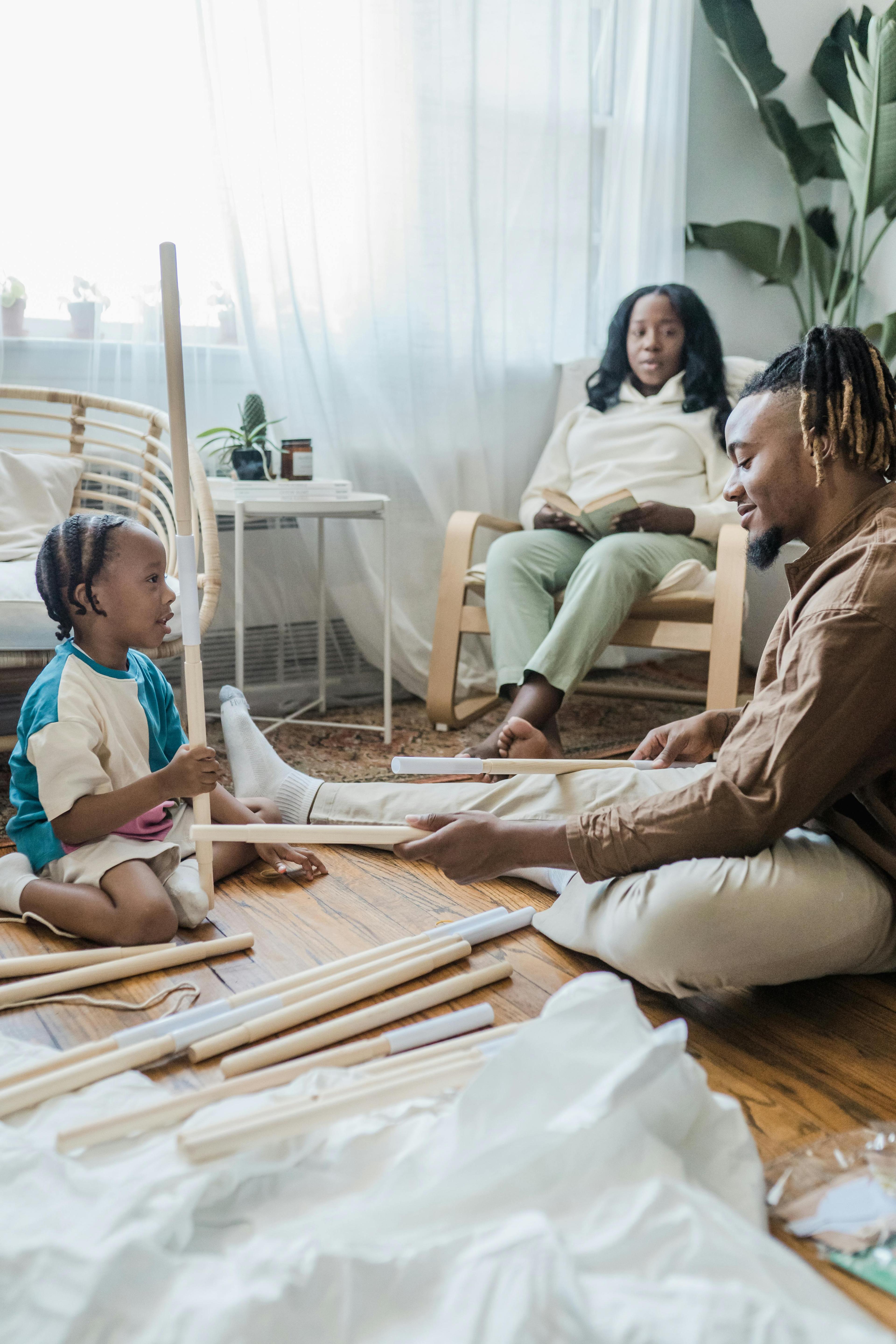 A child and a man sitting on the floor holding some sticks. There is also a woman sitting in a chair with a book in her hand thinking about if she should break up.