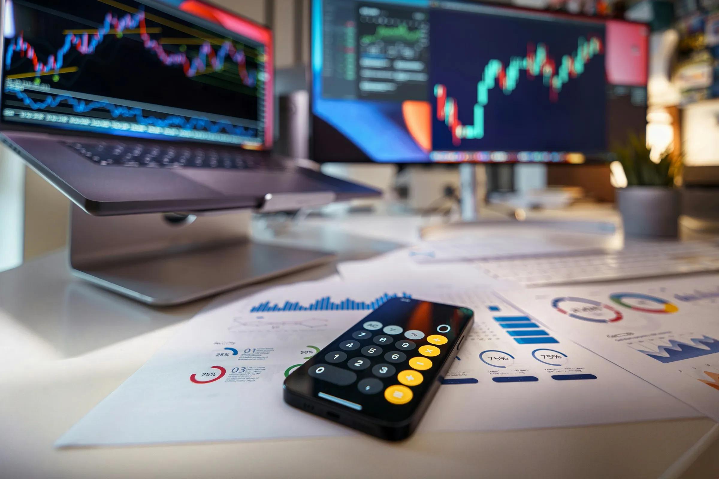Financial market charts displayed on computer monitors above printed trading reports and a smartphone on a desk