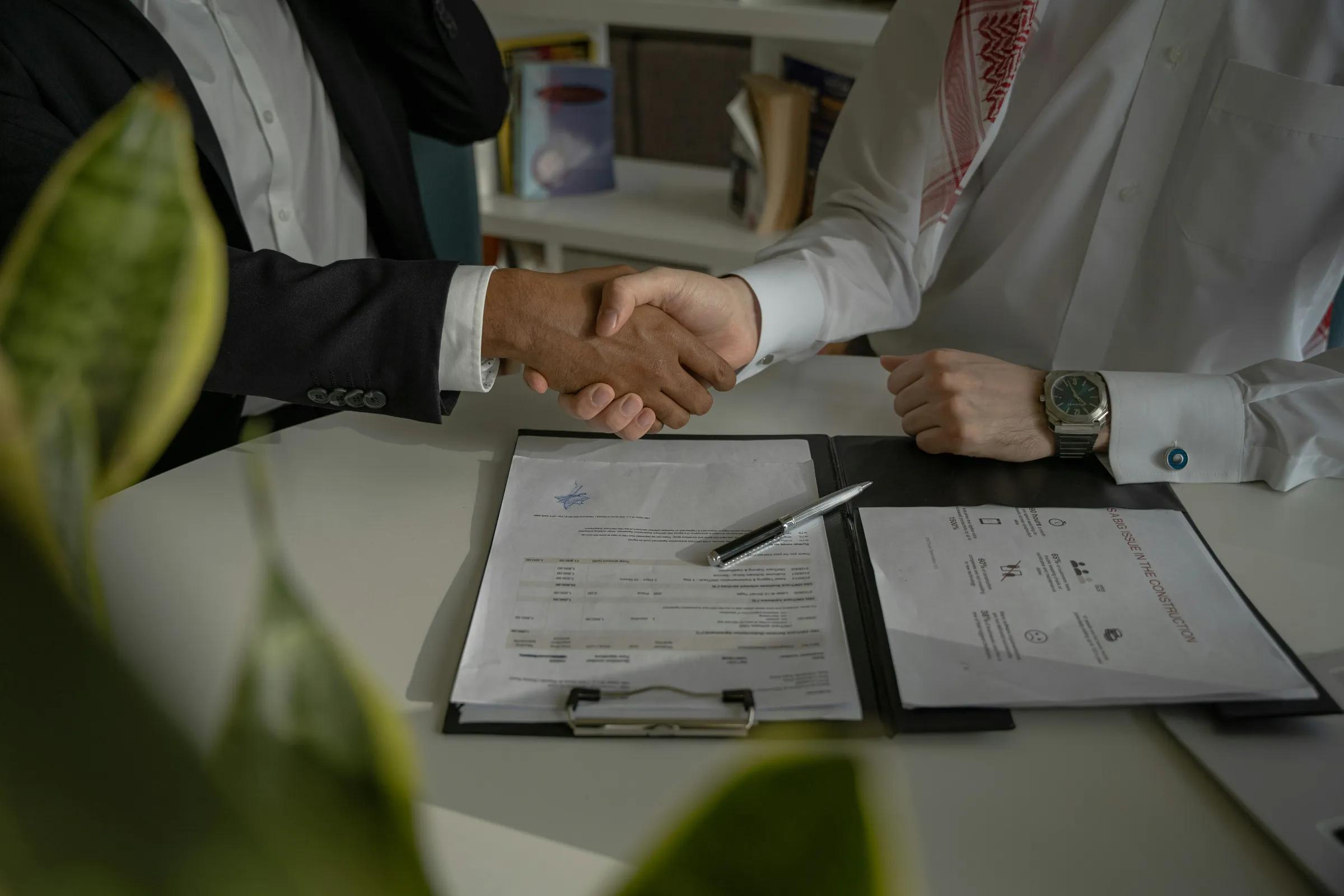 Two business professionals shaking hands over signed contracts on a desk, finalizing a formal business agreement.