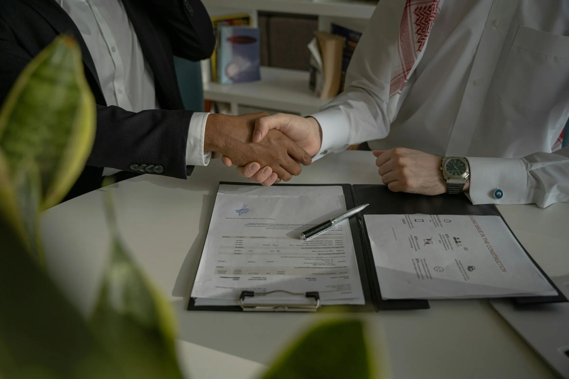 Two business professionals shaking hands over signed contracts on a desk, finalizing a formal business agreement.