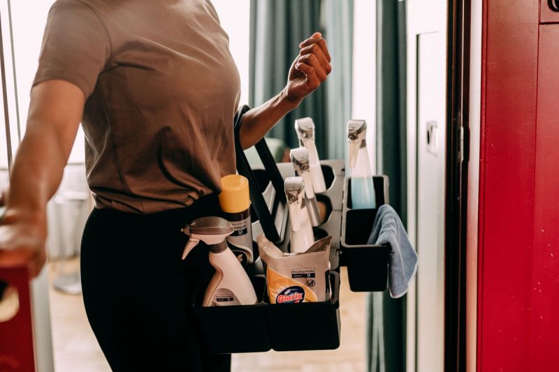 A person carries a black cleaning caddy filled with spray bottles, a can, and a blue cloth.