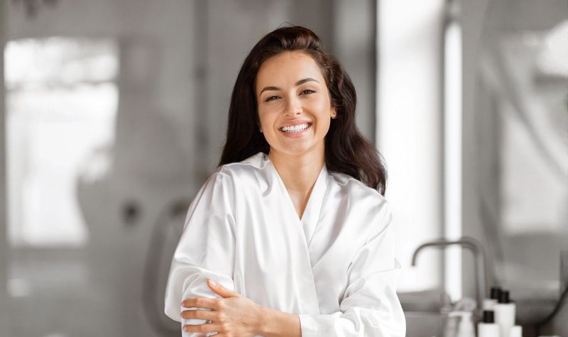 Smiling woman in a white satin robe in a bright bathroom.