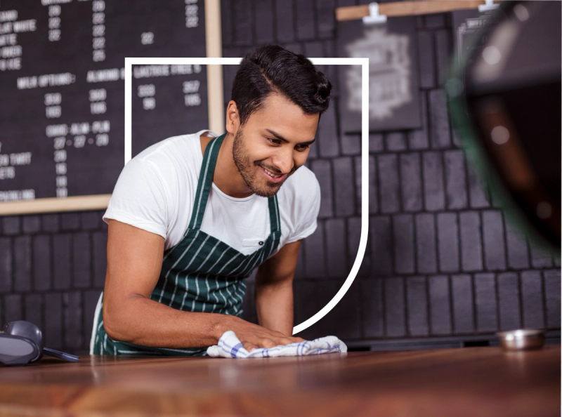 A smiling young man wearing a white t-shirt and green and white striped apron is wiping down a wooden surface, likely cleaning in preparation for opening a cafe or restaurant.