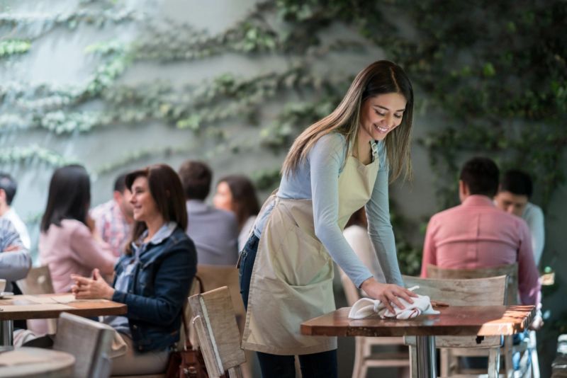 A smiling waitress wearing a beige apron and light blue long-sleeve shirt wipes down a table at a busy cafe.