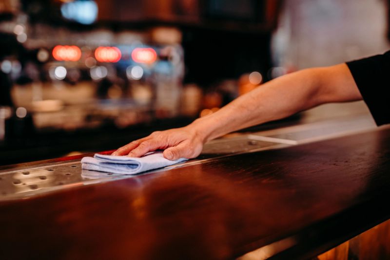 The hands of a bartender or server wiping down a polished wooden bar counter in a dimly lit restaurant or bar setting, with blurred colorful lighting and glassware visible in the background.
