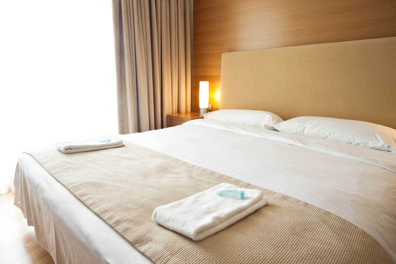 A neatly made hotel bed with crisp white linens, beige runner, and fresh white towels placed on top. A warm bedside lamp glows against the wooden wall panel in the background, and grey curtains frame the side.
