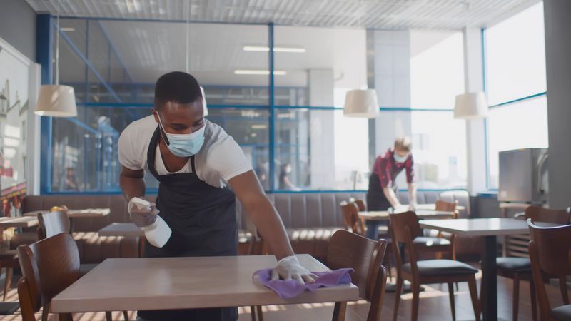 A man wearing a face mask and an apron wiping down a table in a restaurant or café.