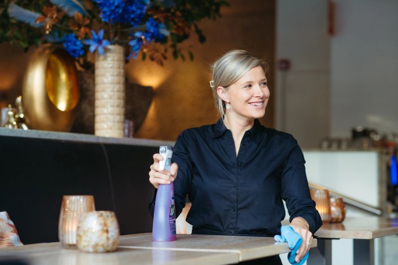 A smiling blonde woman in a black shirt cleans a restaurant table with a spray bottle and blue cloth.