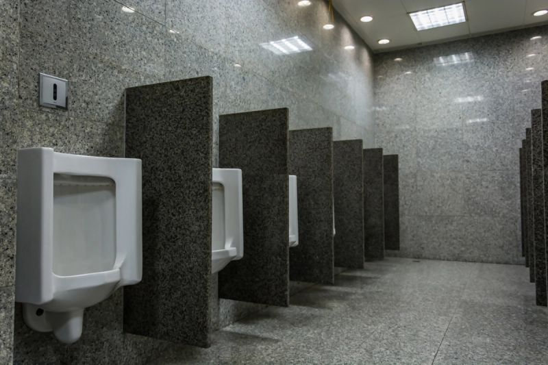 The interior of a public toilet or restroom with walls and floor covered in tiles of granite or similar stone material. There is a row of urinals, each unit separated by granite dividers. The room has a spotlessly clean and well-maintained appearance.