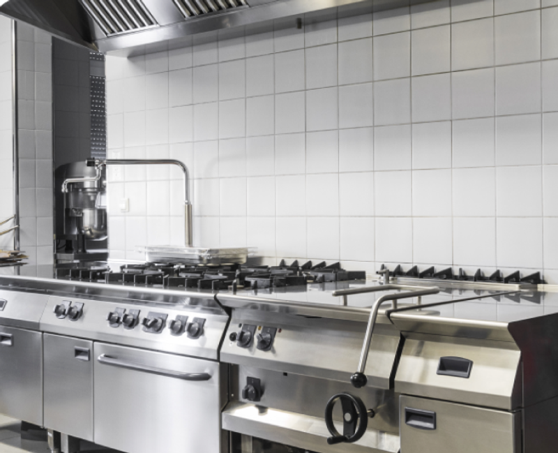 A professional commercial kitchen with tiled walls and featuring a large stainless steel gas stove with multiple burners and an extractor hood above. The equipment appears to be well-maintained and organized, suggesting a focus on efficiency and cleanliness in the kitchen.