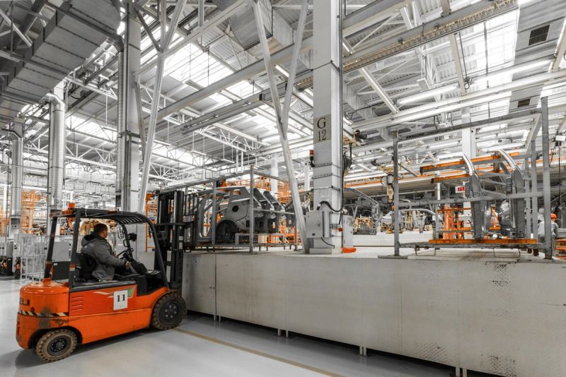 The interior of a large industrial manufacturing facility or warehouse. The space features high ceilings with exposed metal trusses and ductwork. There are various pieces of industrial machinery and equipment visible, including an orange forklift truck being operated by a worker.