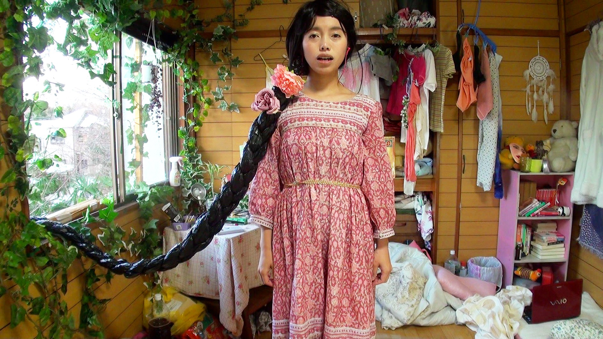 A person in a pink patterned dress standing in a cluttered bedroom, with an exaggerated long black braid extending across the room.