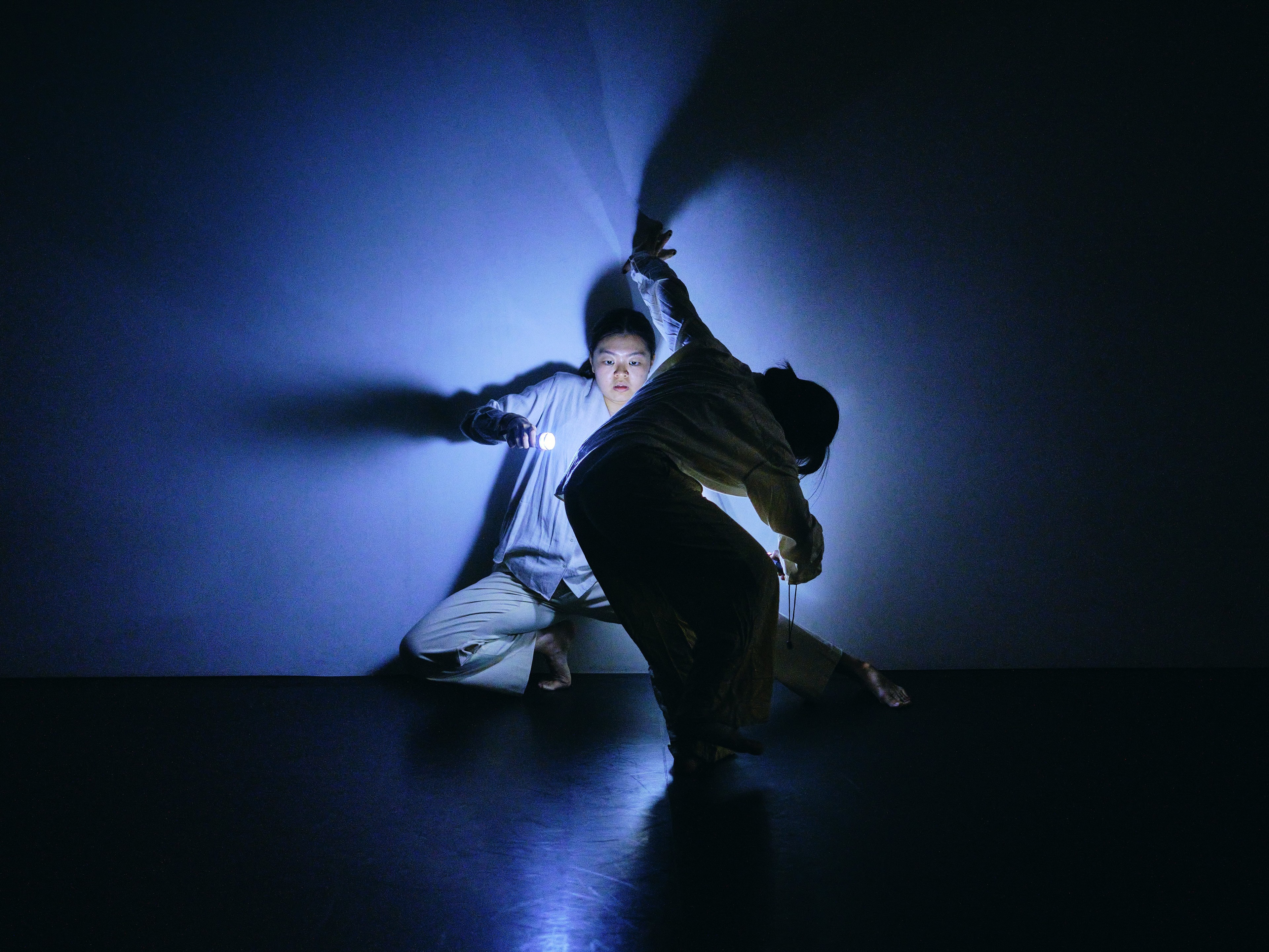 Two performers moving together in a dark space, dramatically illuminated by blue light that casts elongated shadows behind them.