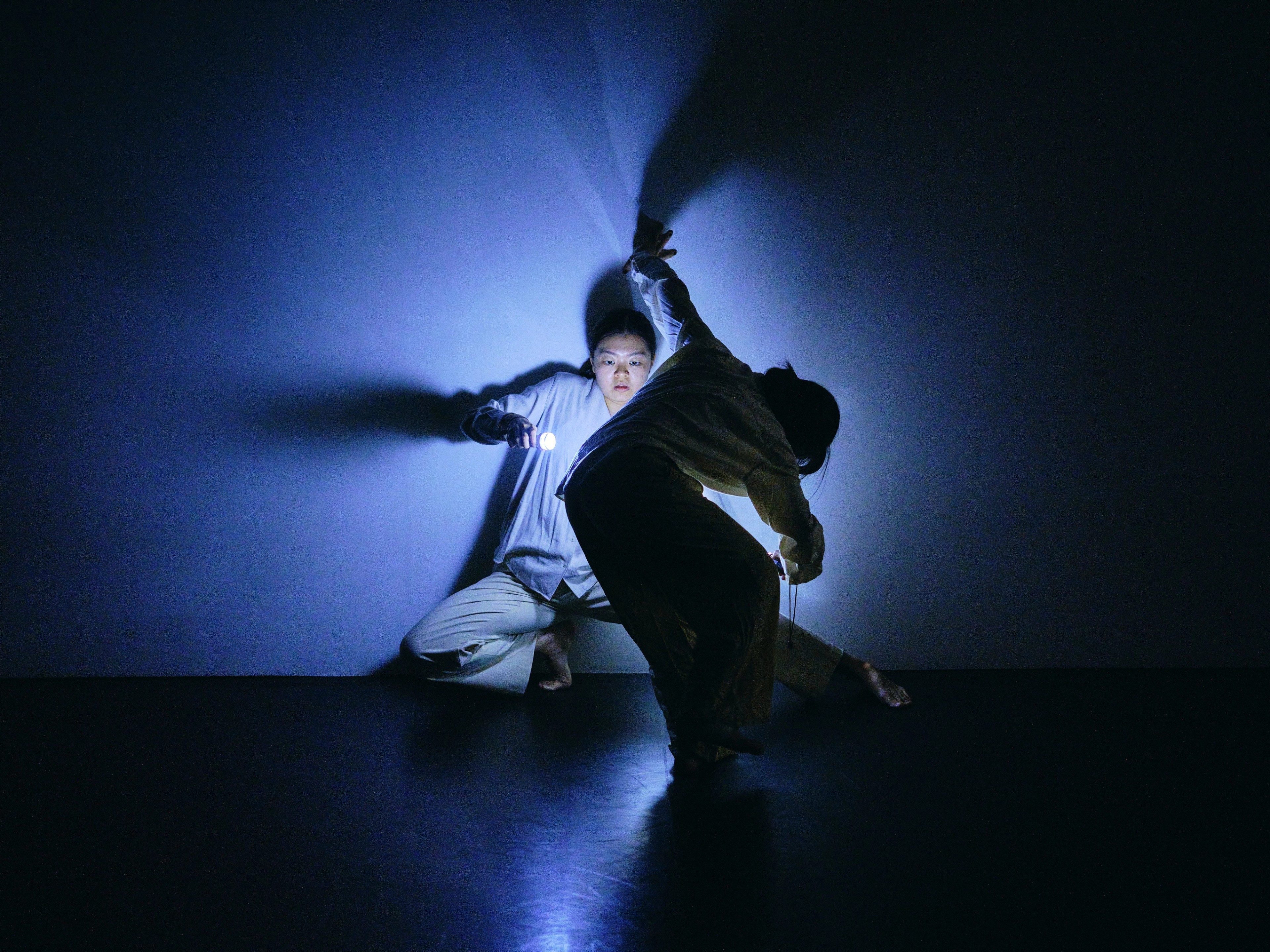 Two performers moving together in a dark space, dramatically illuminated by blue light that casts elongated shadows behind them.