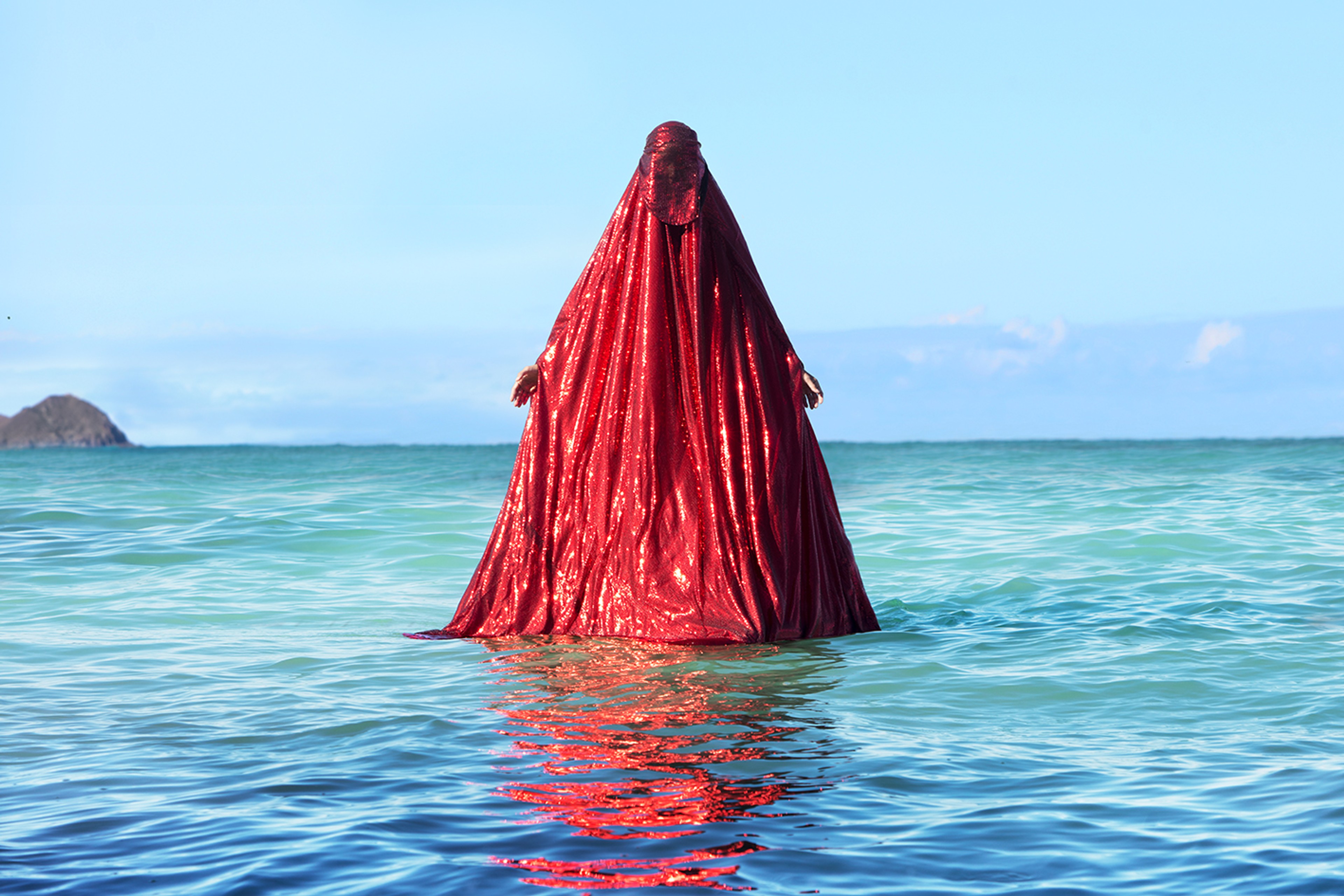 A figure completely draped in shimmering red fabric standing waist-deep in the sea under a clear blue sky.
