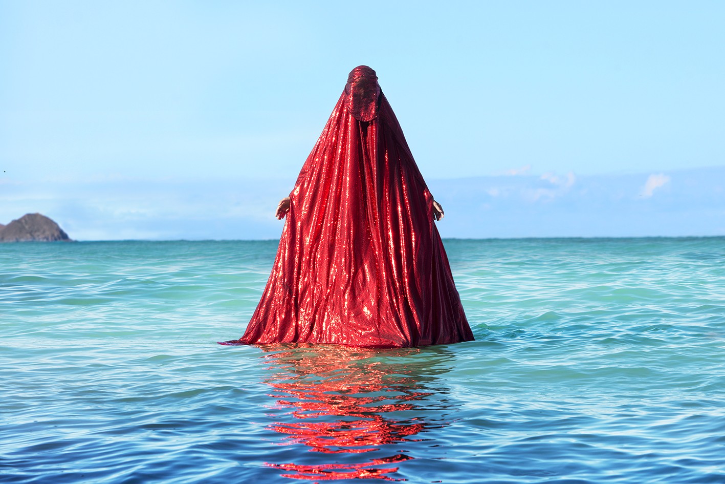 A figure completely draped in shimmering red fabric standing waist-deep in the sea under a clear blue sky.