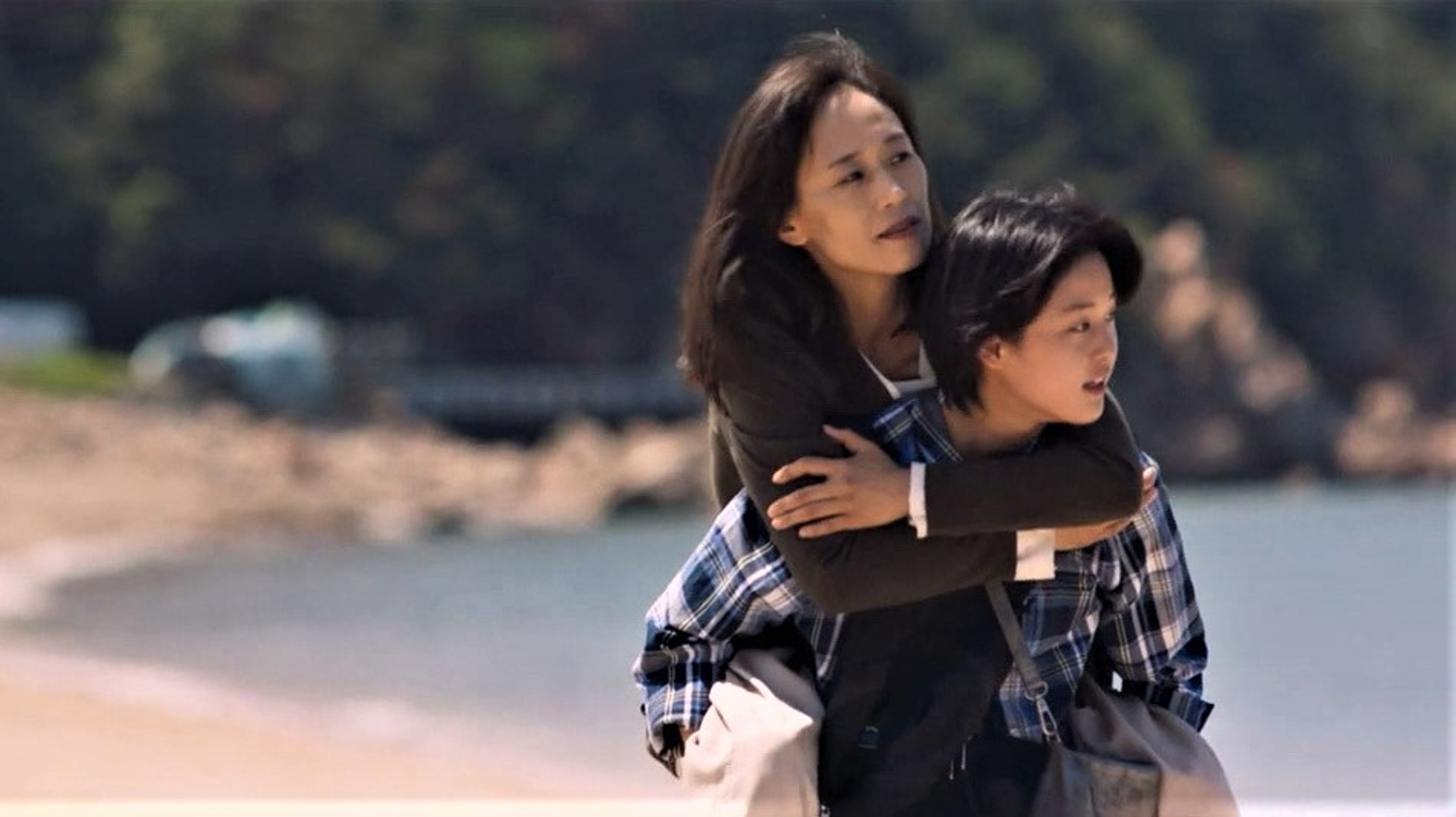 One woman carrying another piggyback along a beach, with the sea and rocky shoreline behind them.