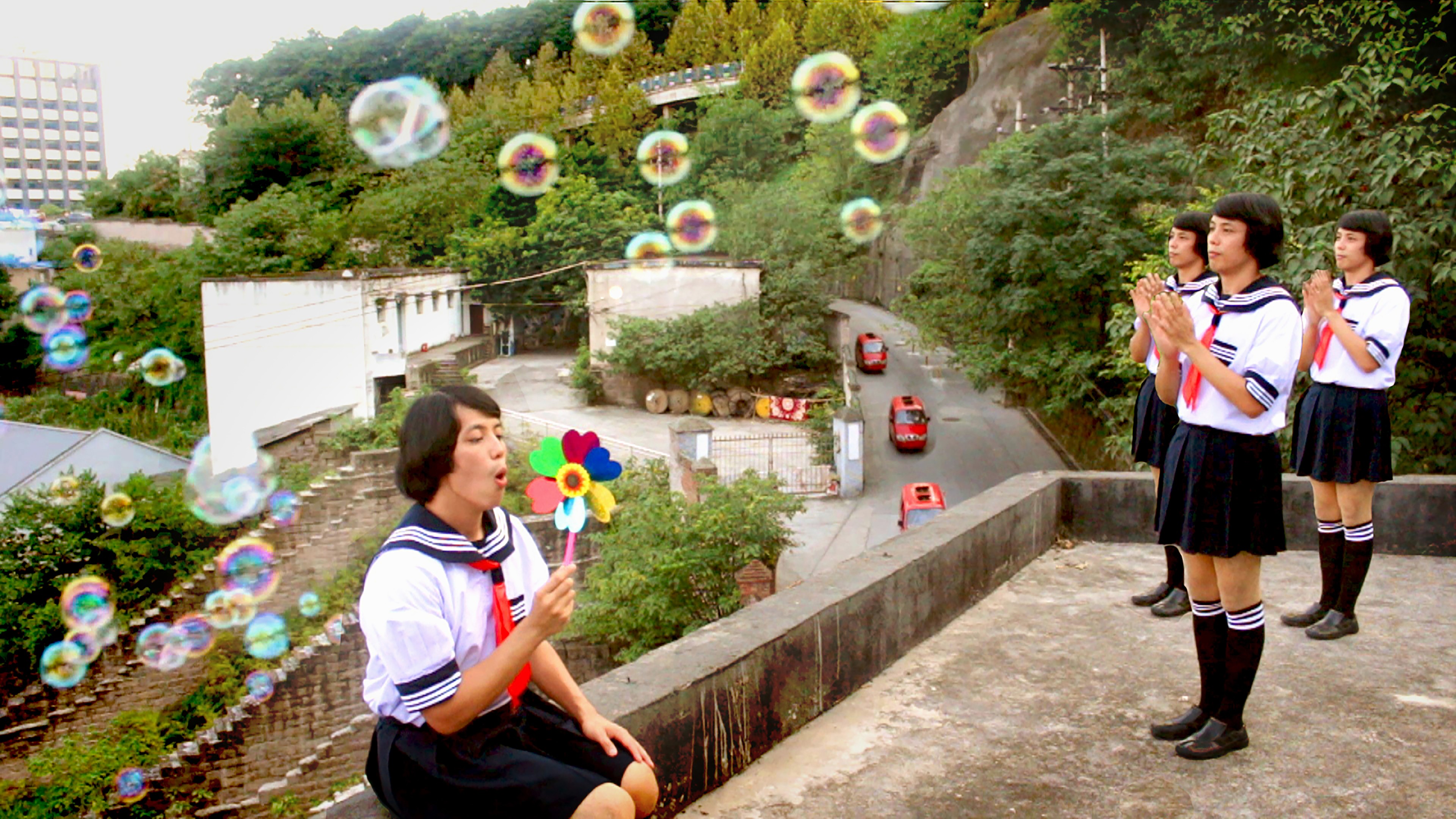 A group of performers in matching school uniforms creating bubbles on a rooftop overlooking a green hillside and city road.