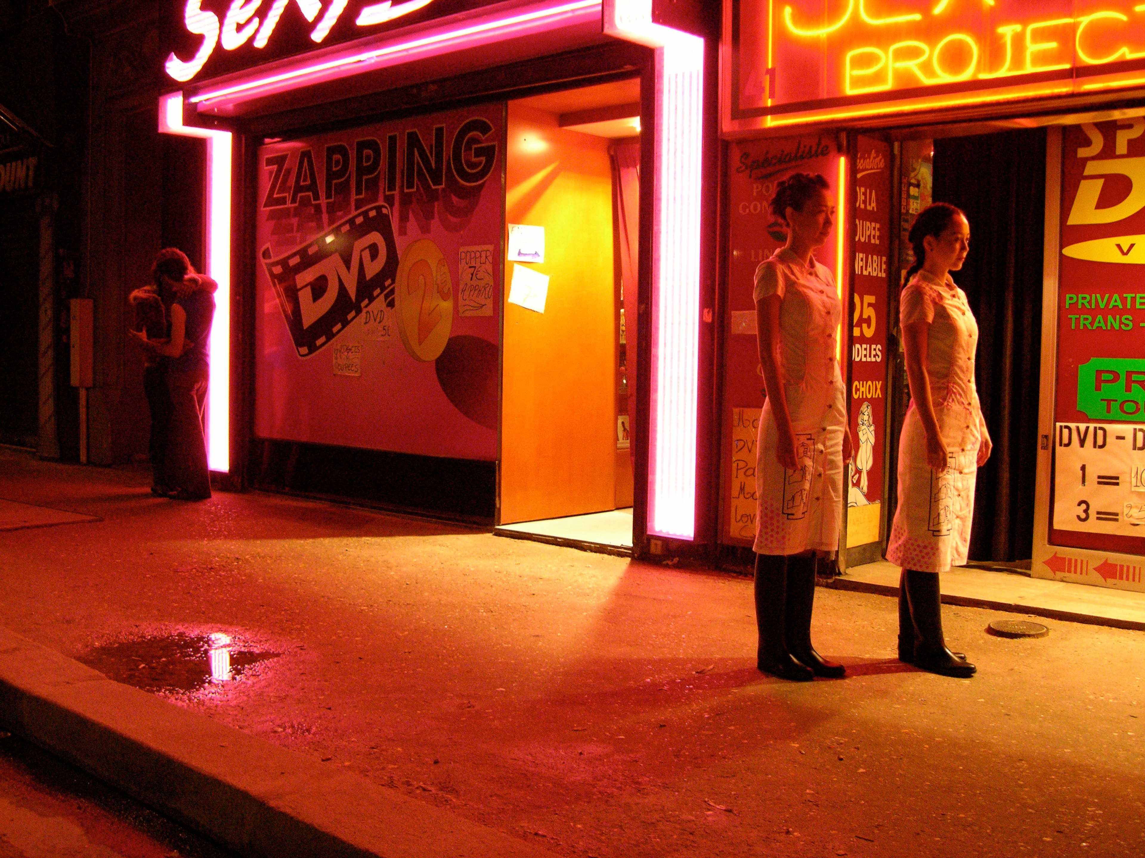 Two people standing outside a brightly lit adult entertainment venue at night, surrounded by neon signage.