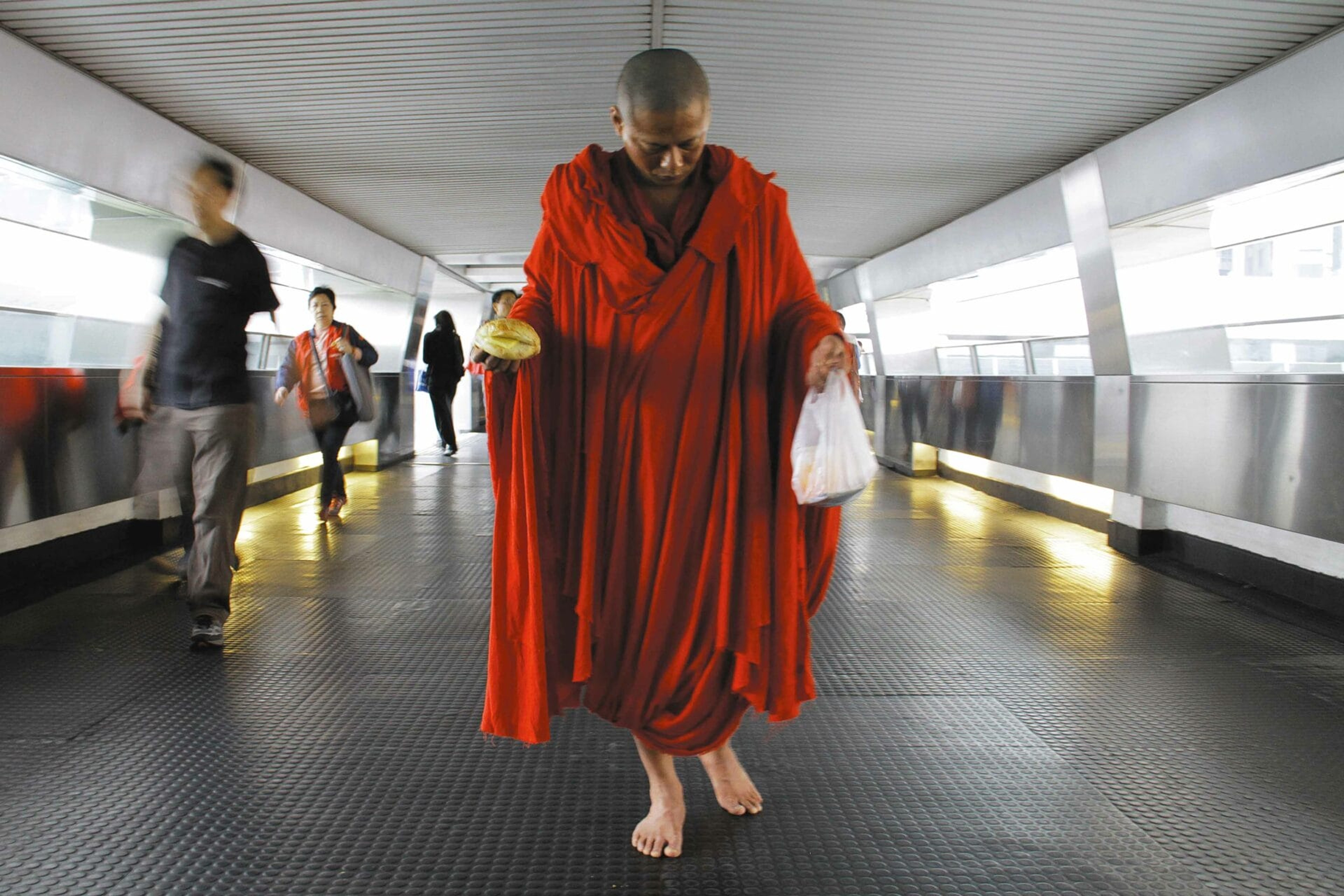 A barefoot person in flowing red robes walking through a modern pedestrian passage as commuters pass by.