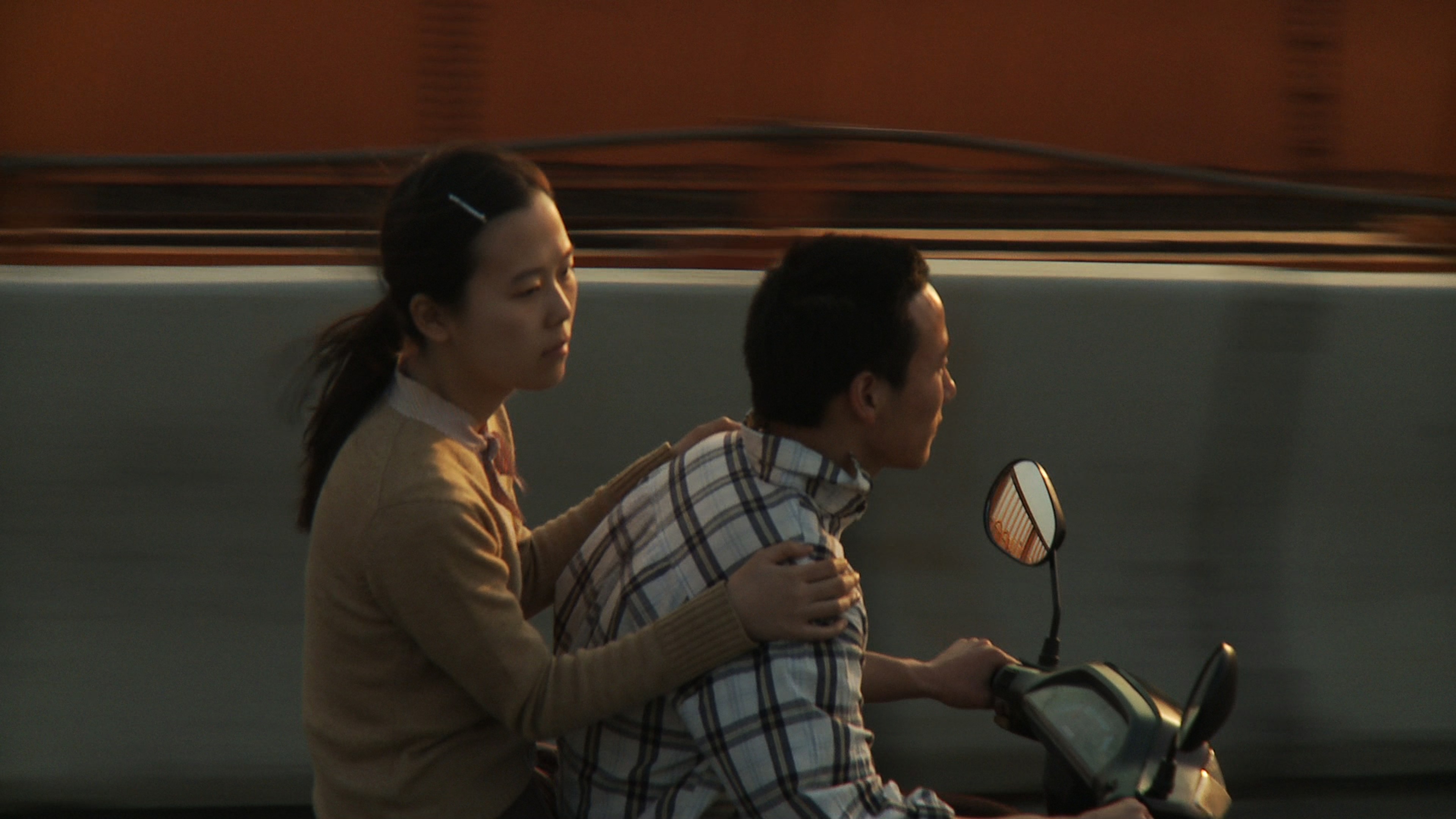 A woman riding as a passenger on a scooter, holding onto the driver as they travel along a road.
