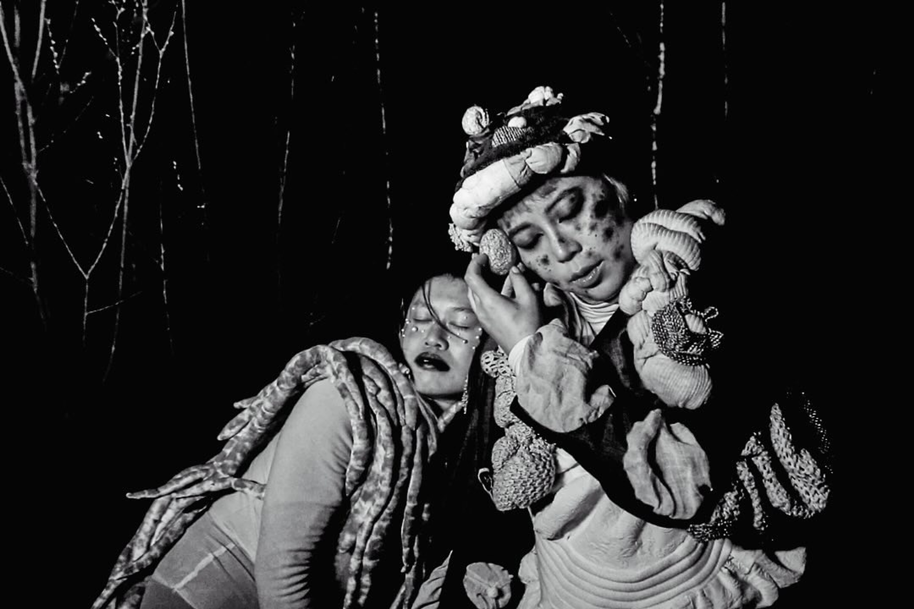 Two elaborately costumed performers leaning against each other in a black-and-white scene.