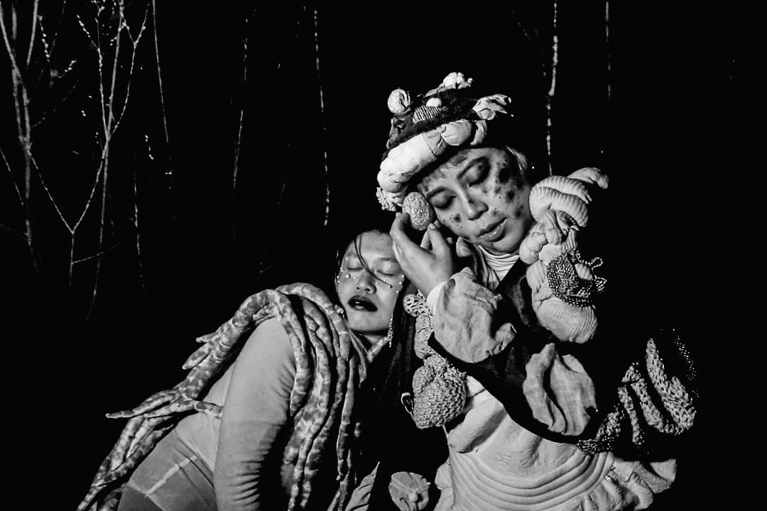 Two elaborately costumed performers leaning against each other in a black-and-white scene.