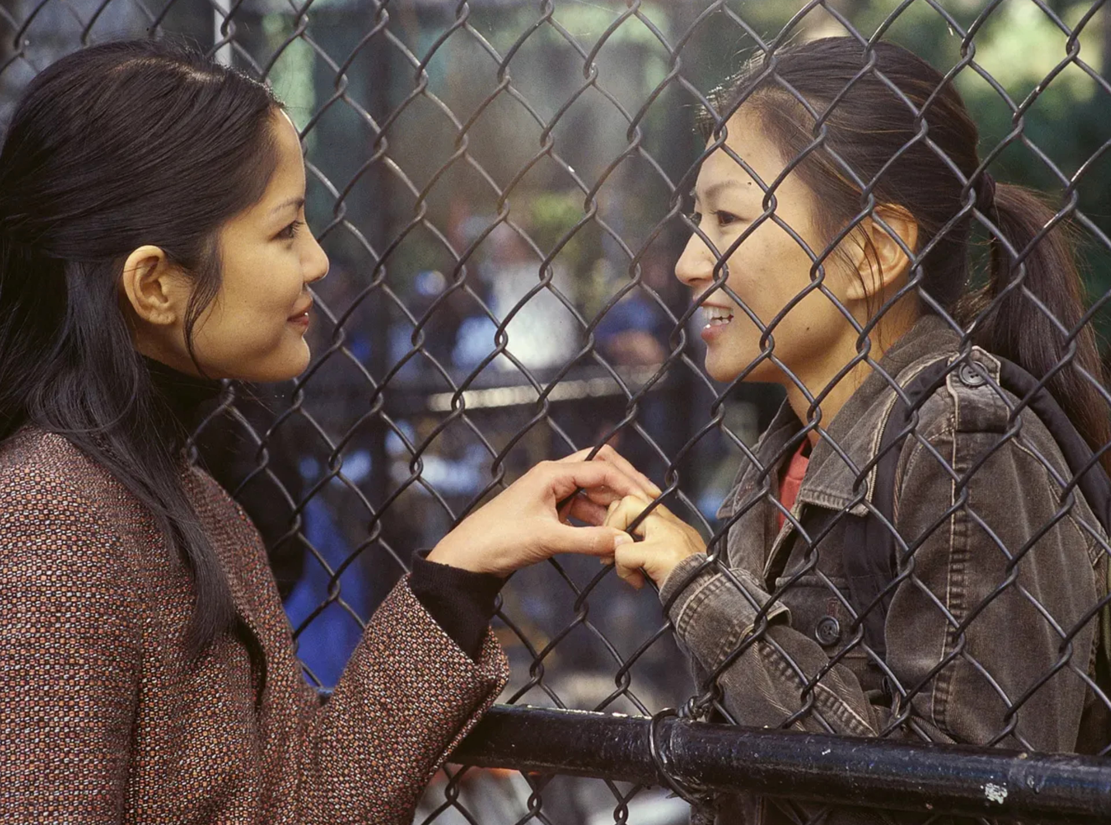 Two women smiling at each other through a chain-link fence, gently touching hands.