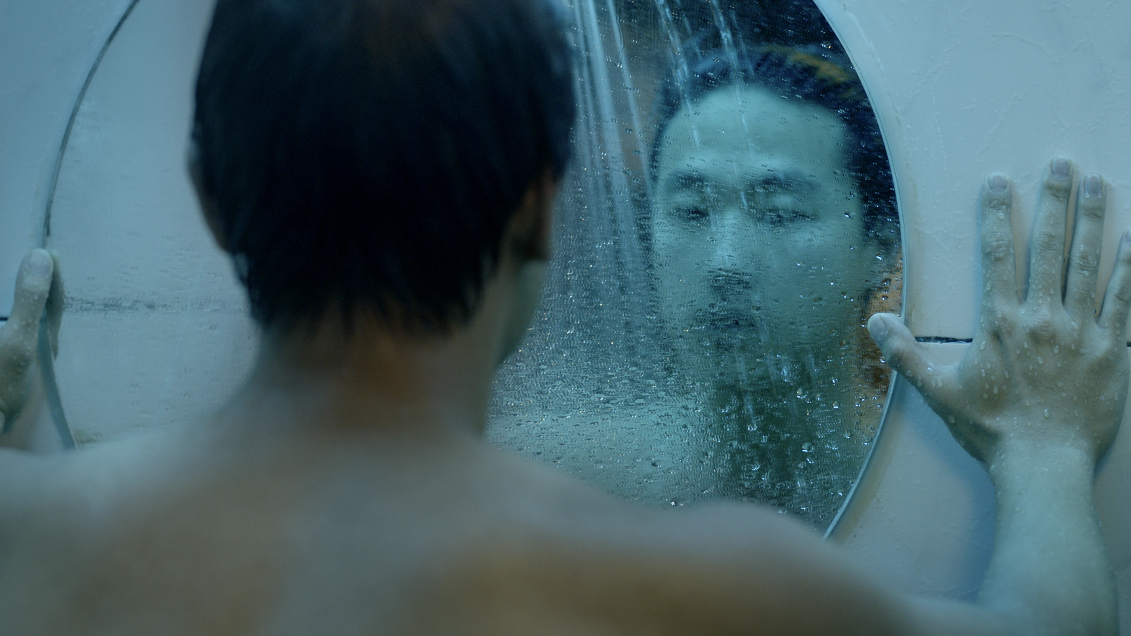 A shirtless person pressing their hands against a fogged circular mirror, their face reflected through the condensation.