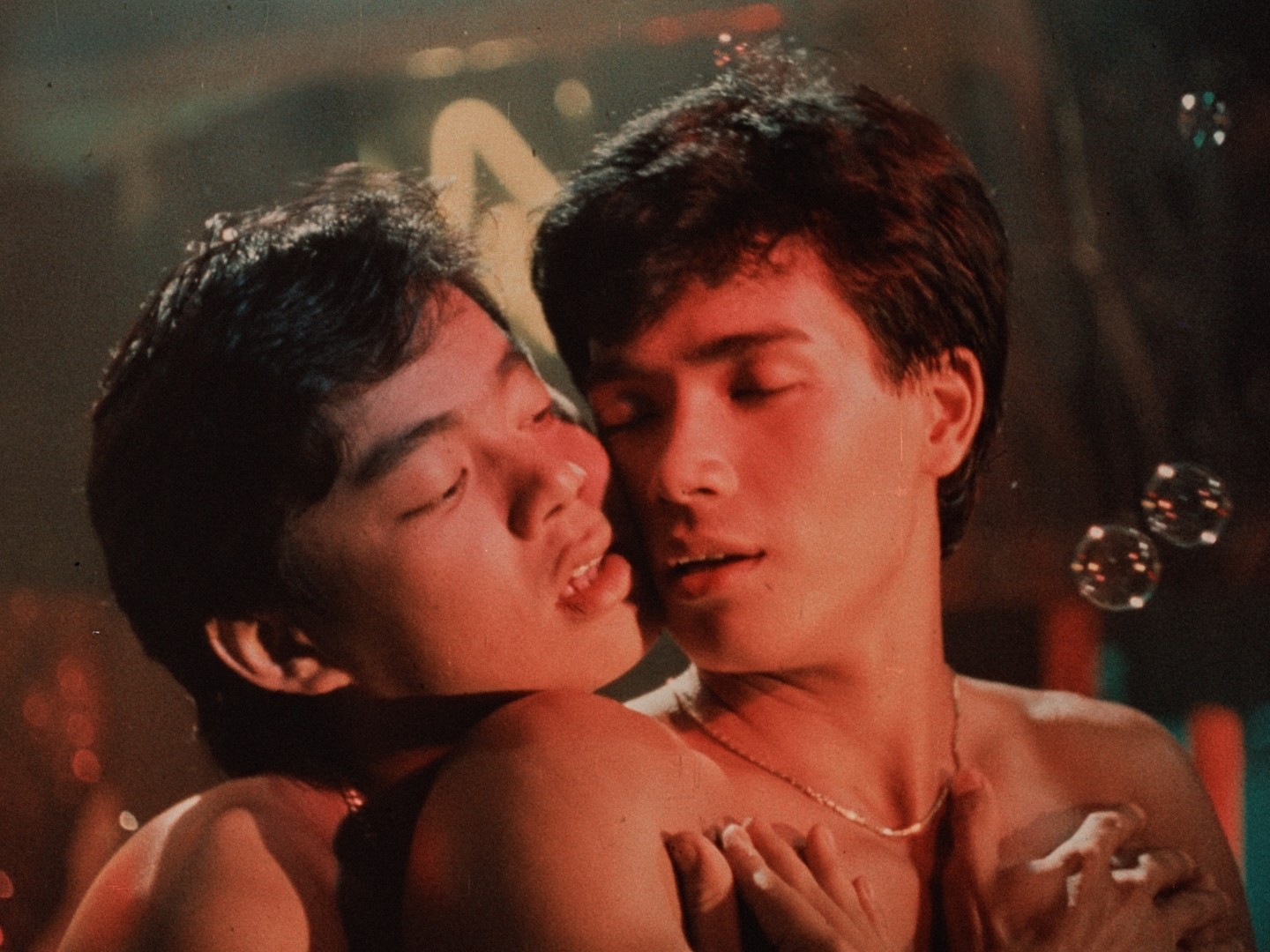 Two shirtless men embracing closely in a dimly lit scene.