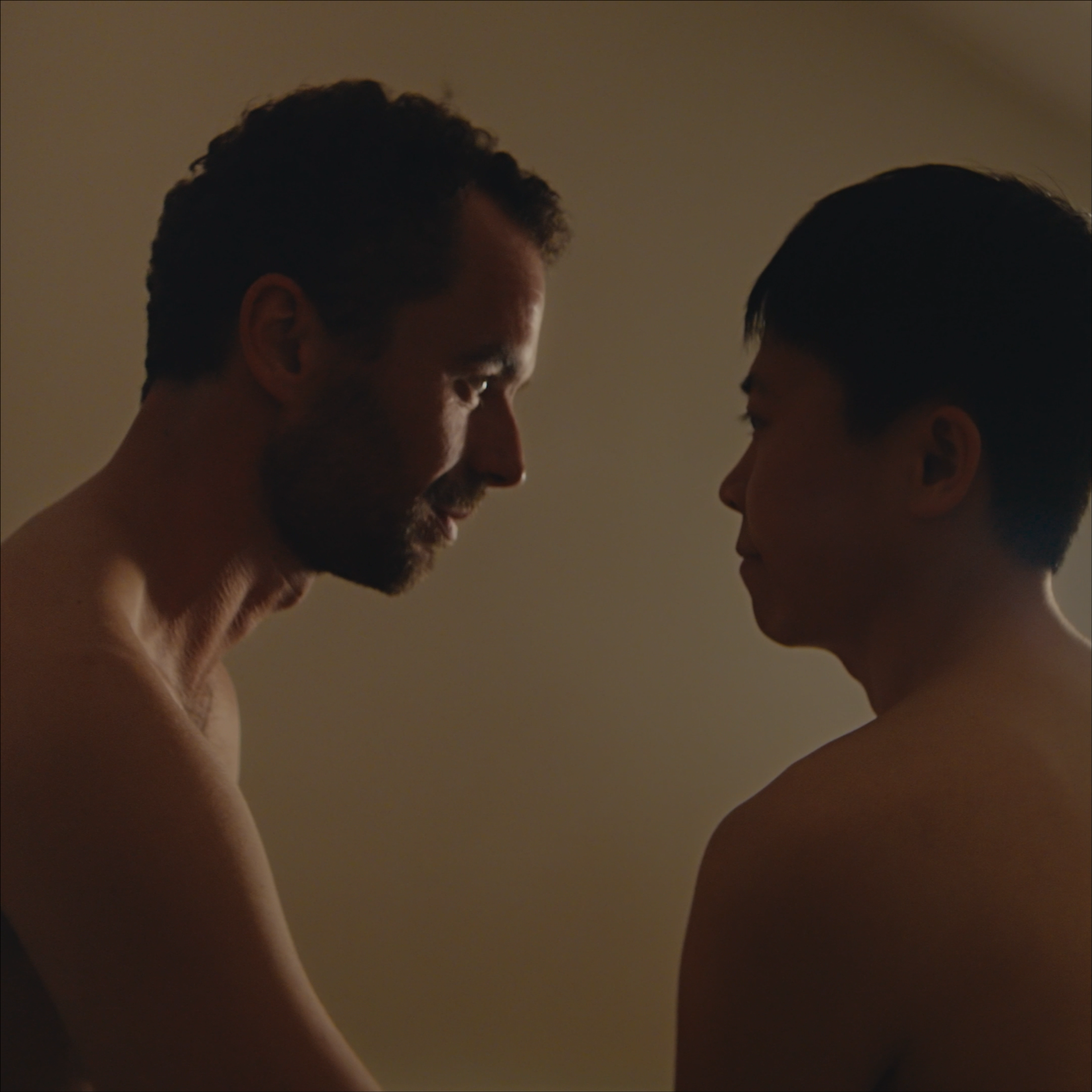 Two shirtless men facing each other closely in a dimly lit room.