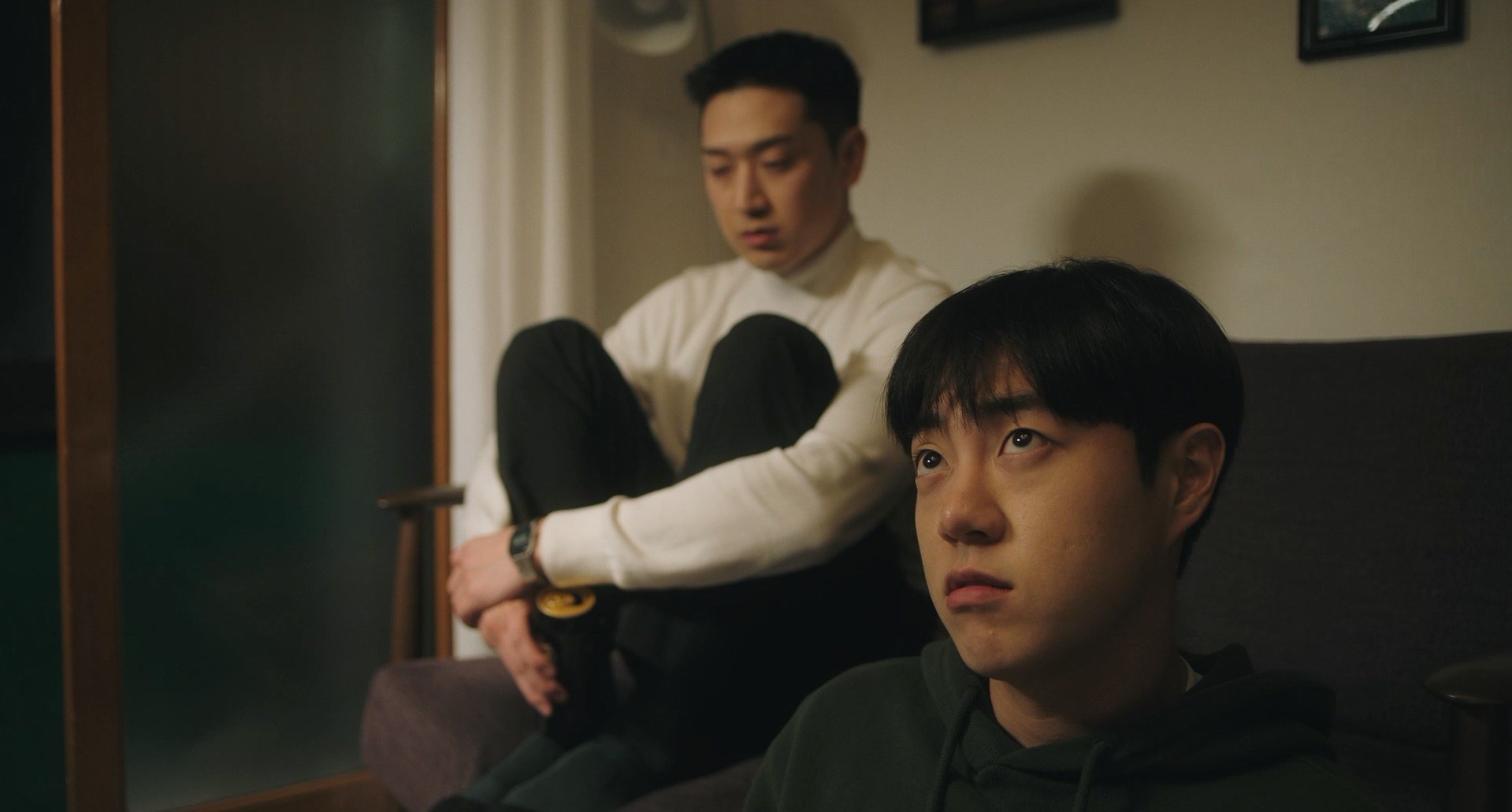 Two young men sitting together in a dimly lit room, one in the foreground looking upward while the other sits behind with his knees drawn up.