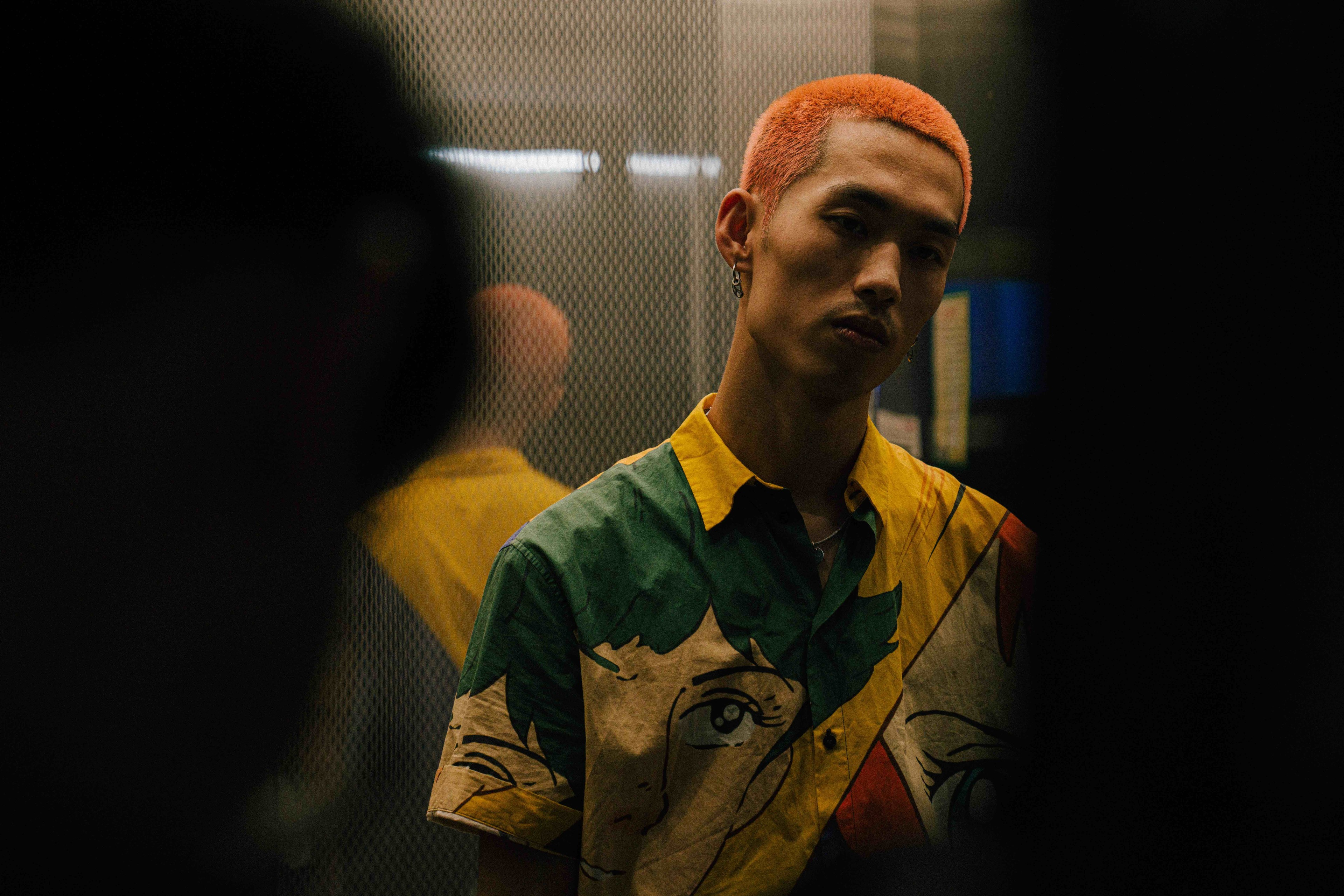 A person with bright orange hair standing in a dim interior, wearing a colourful patterned shirt.