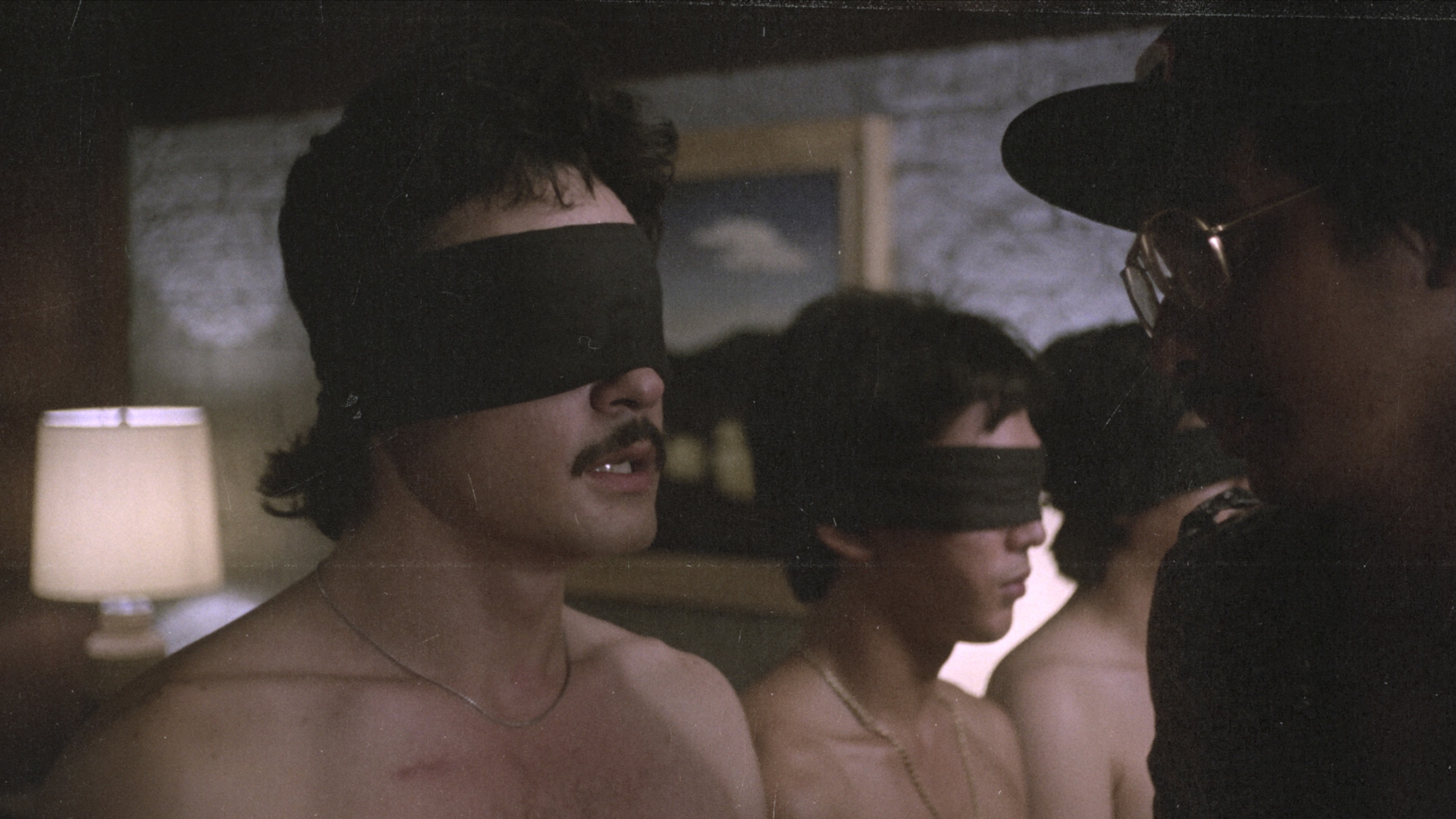 Two shirtless men wearing blindfolds standing closely together in a dimly lit room.