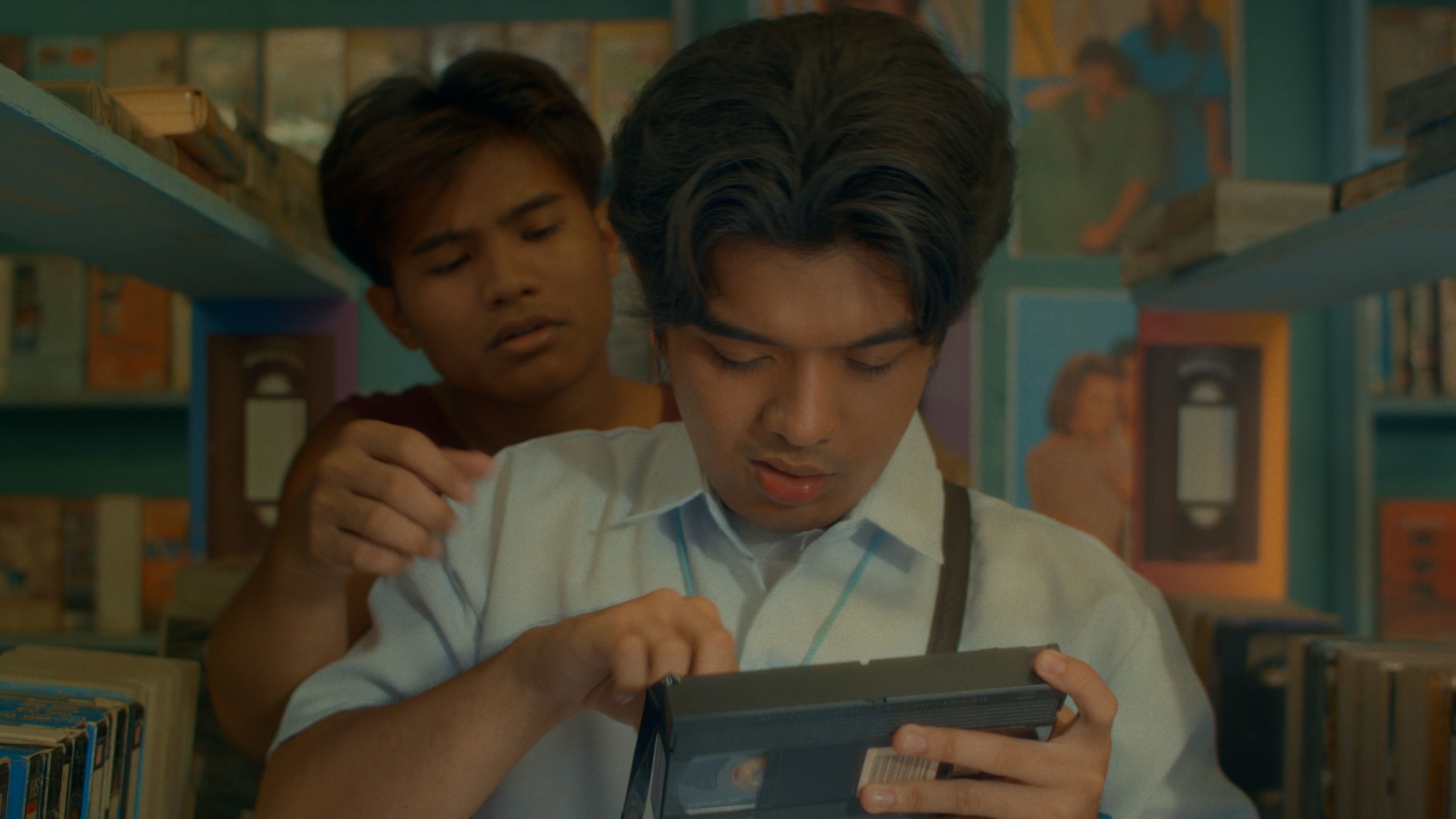 Two young men standing closely together in a video shop, one holding a VHS tape.