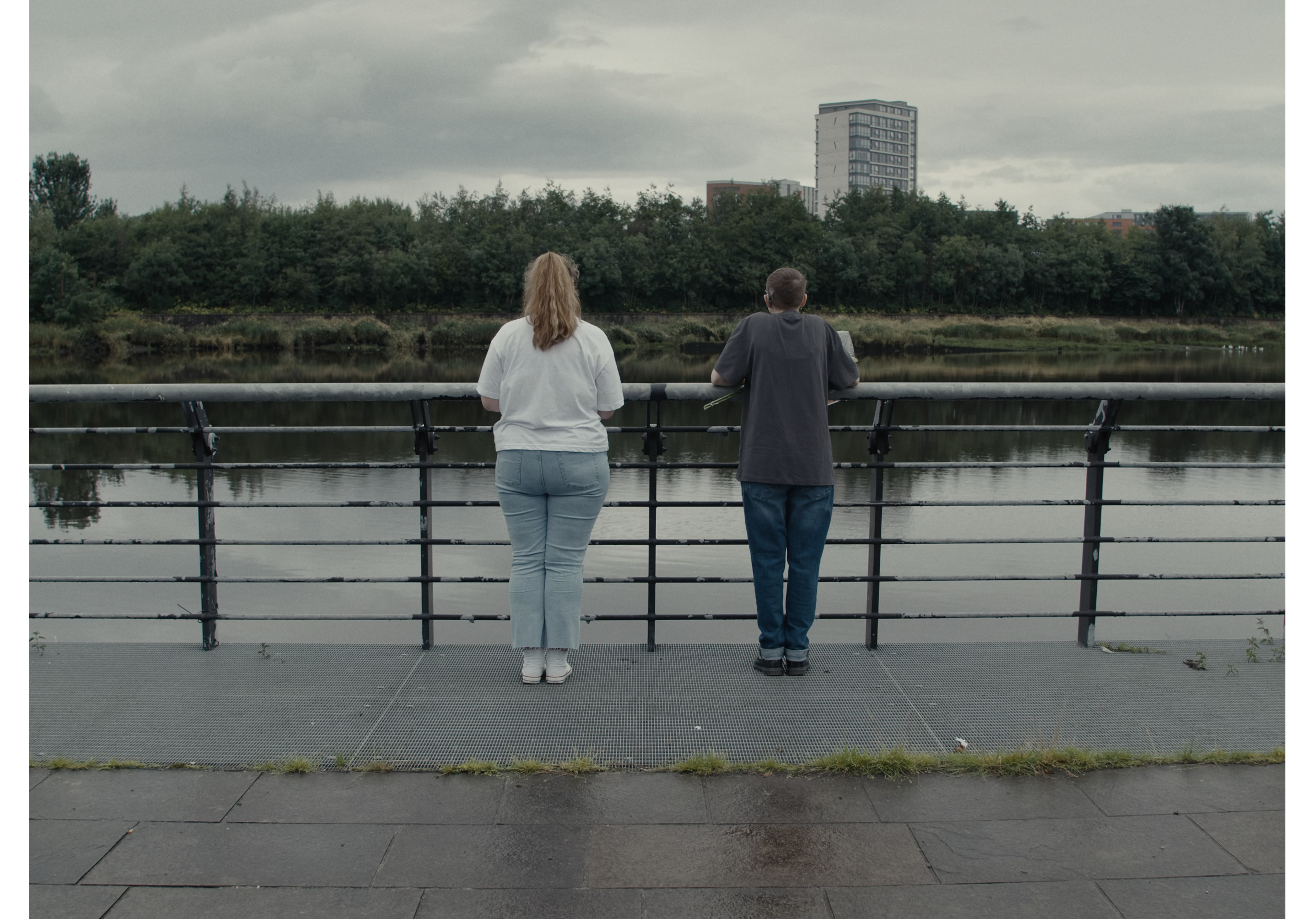Two people standing side by side at a riverside railing, looking across the water beneath an overcast sky.