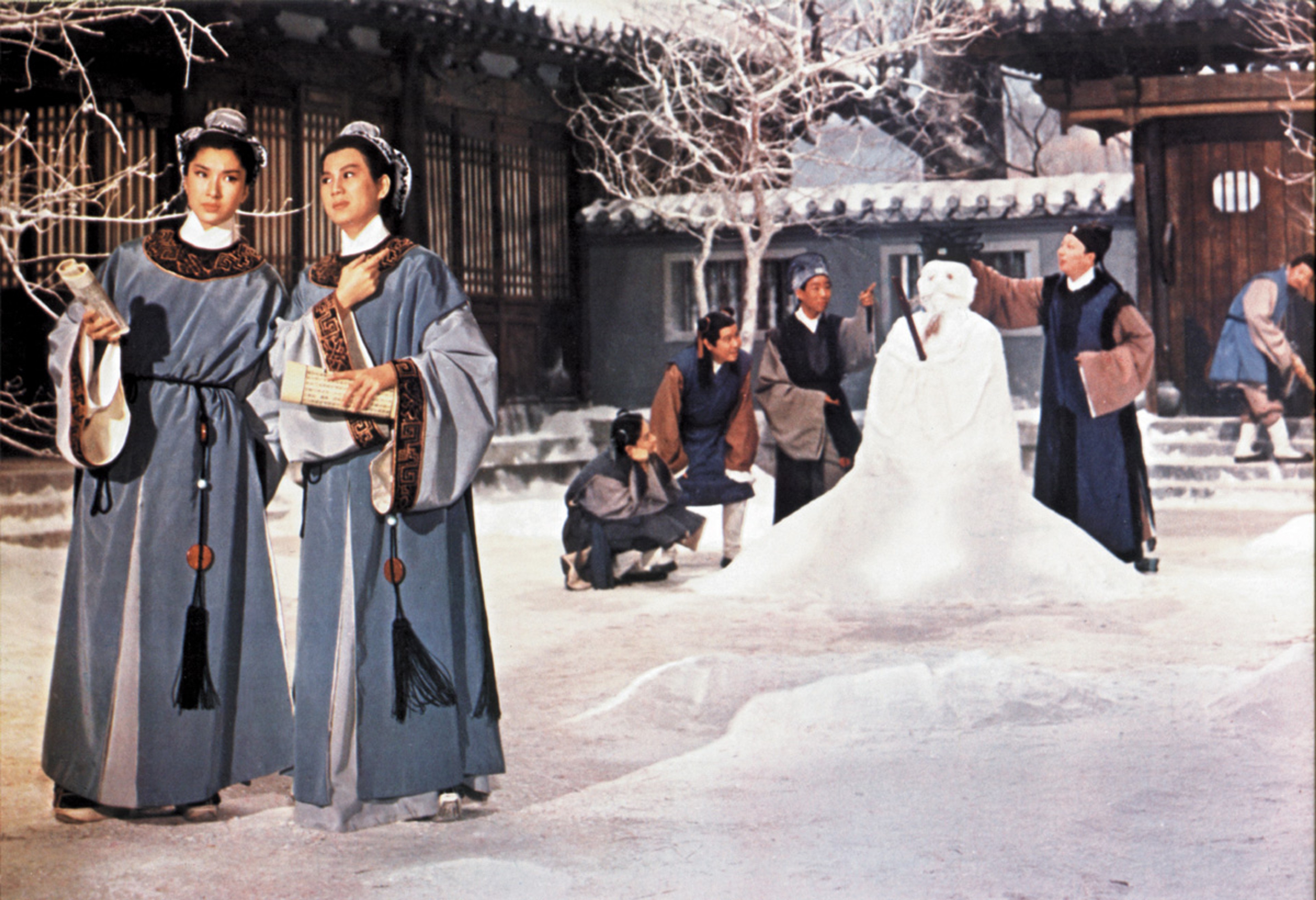 Two women in traditional dress standing in a snowy courtyard, with a large snow sculpture and several people in the background.