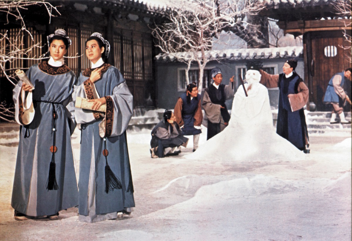 Two women in traditional dress standing in a snowy courtyard, with a large snow sculpture and several people in the background.