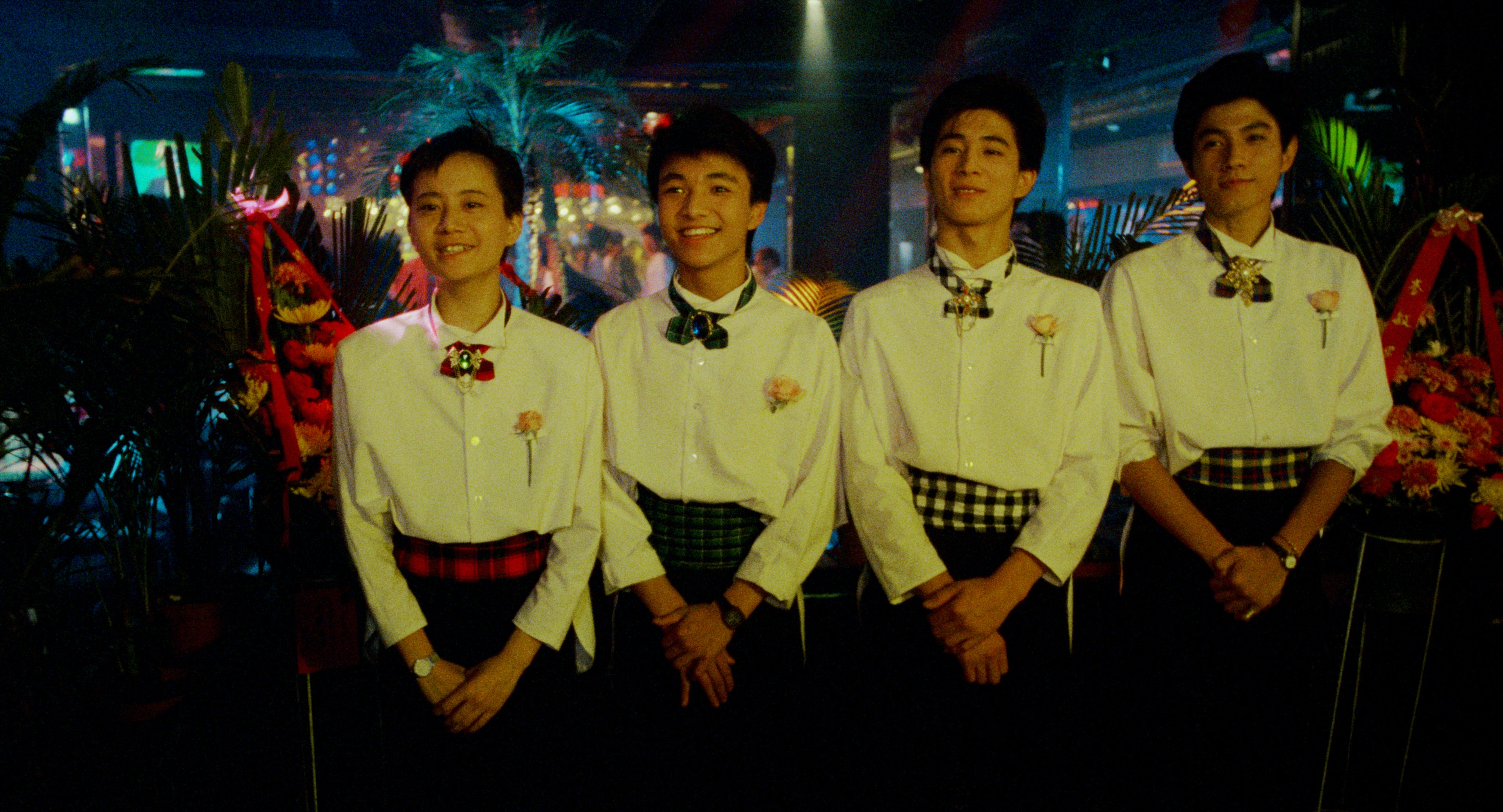 Four people in matching white shirts and bow ties standing together in a colourful nightclub setting.