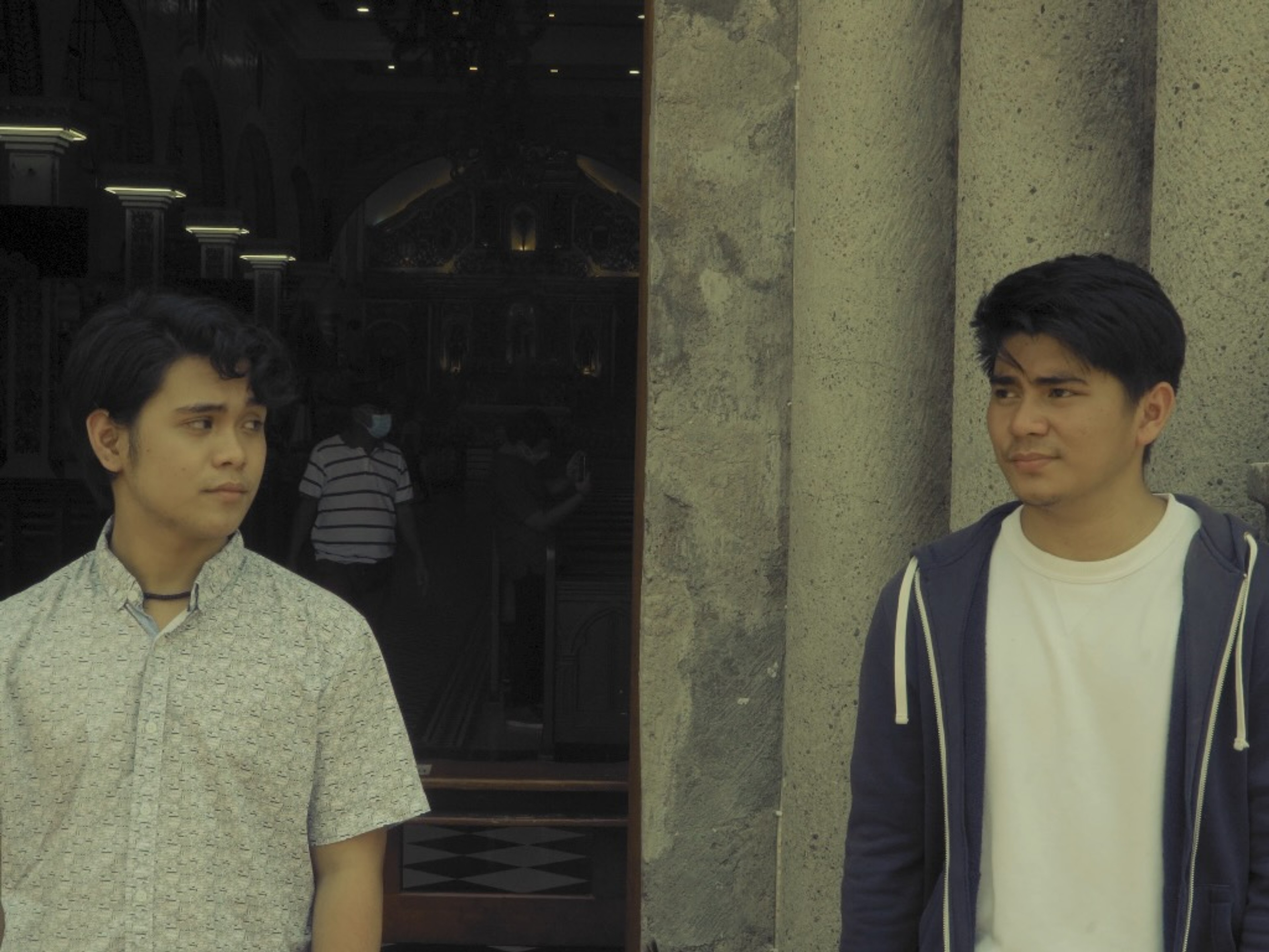Two young men standing apart outside a stone building, looking toward each other.