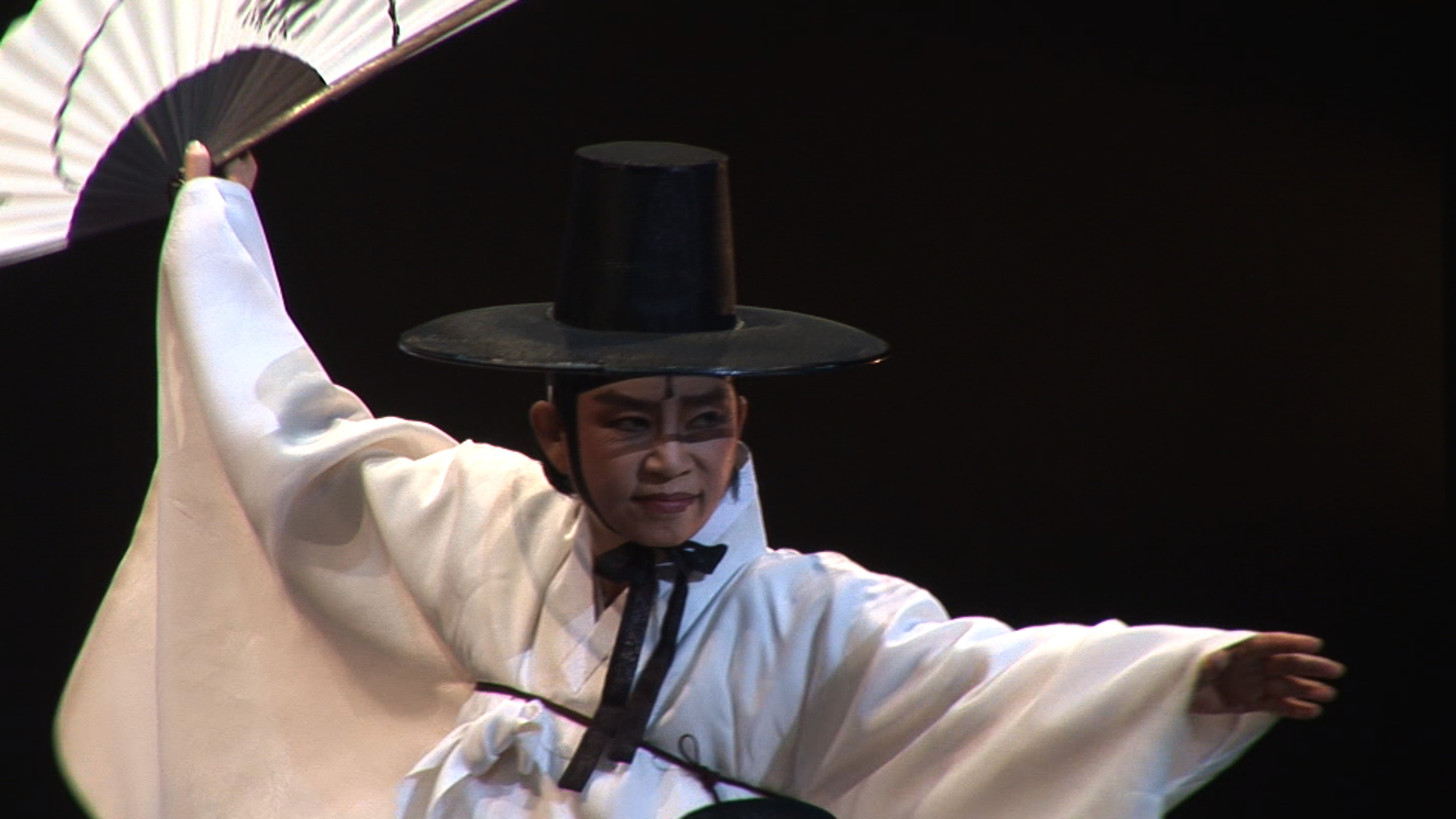 A performer in traditional Korean dress and a black hat dancing with a large fan on stage.