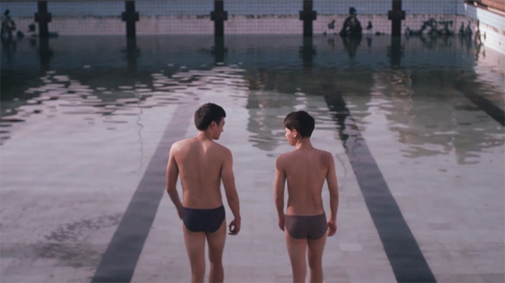 Two young men in swim briefs standing side by side at the edge of a swimming pool, looking toward each other.