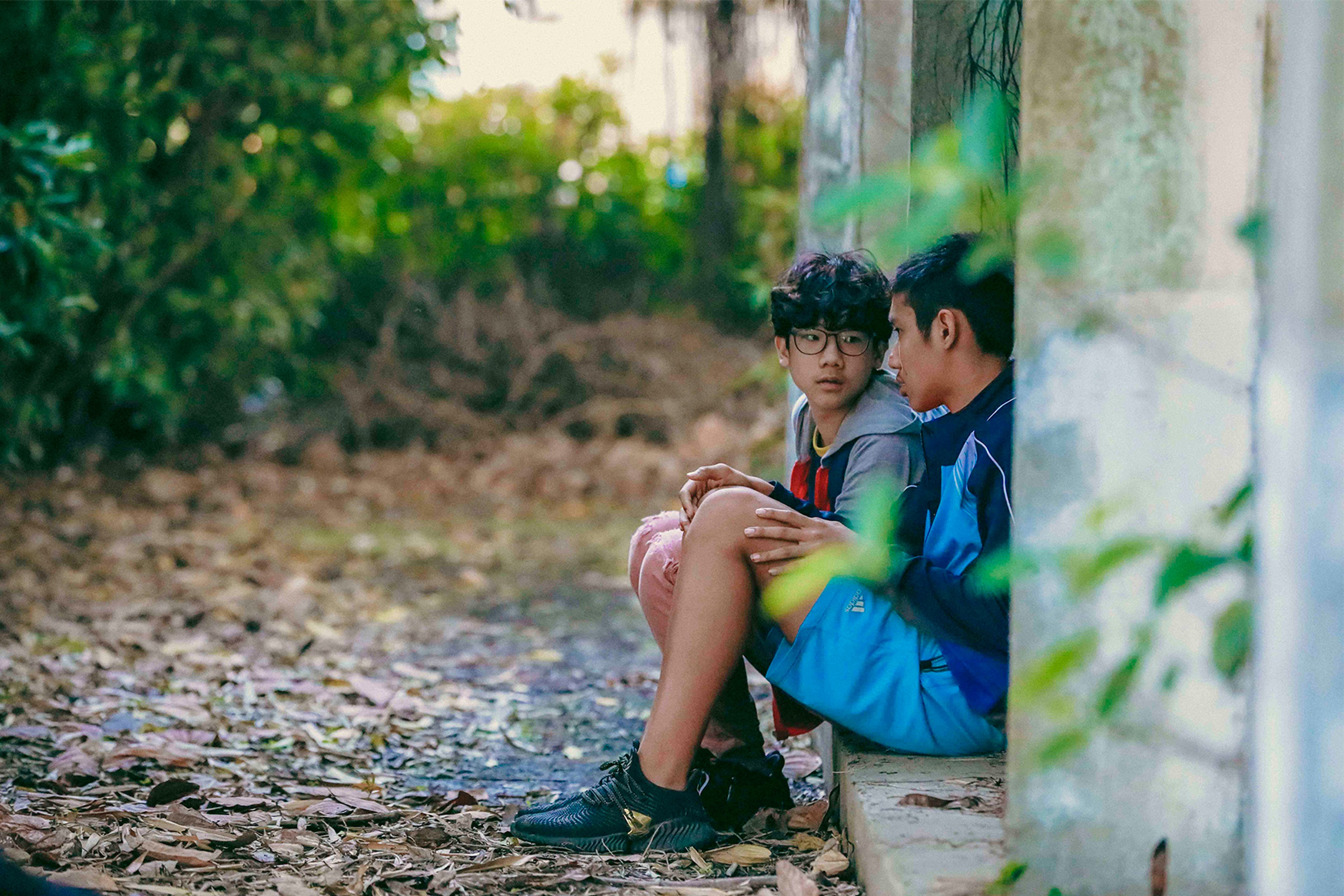 Two young people in sportswear sitting closely together beside a wall in a wooded area.