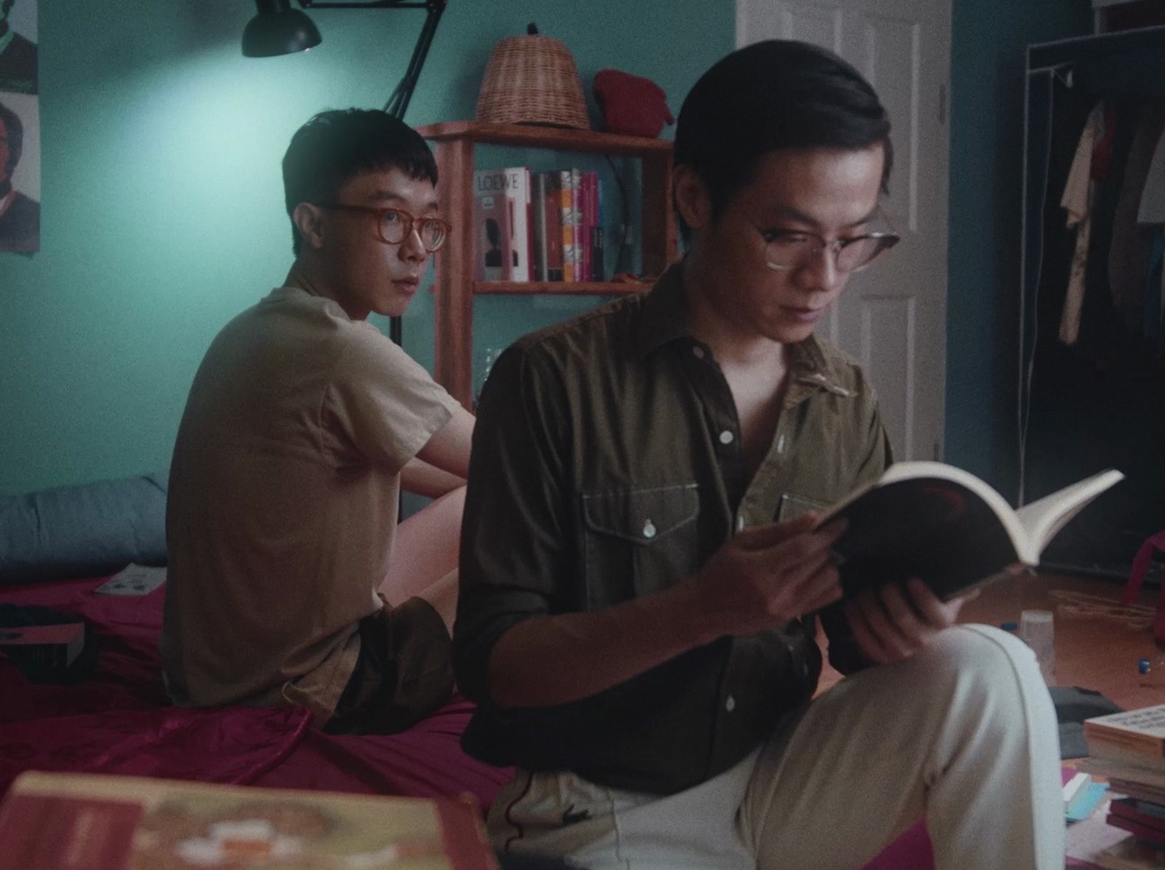 A man in the foreground reads a book while the man in the background looks at him.