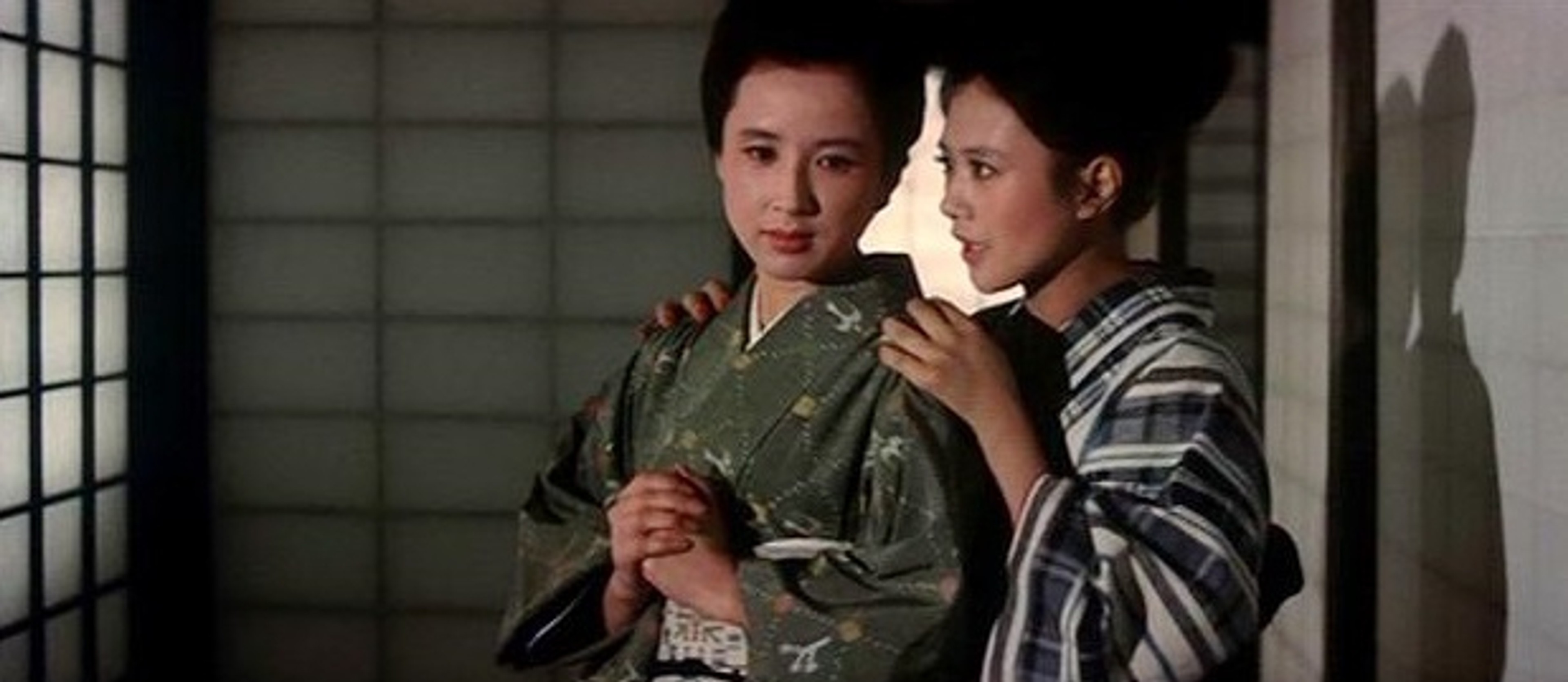 Two women in traditional Japanese dress standing closely together indoors, one resting her hands on the other’s shoulders.