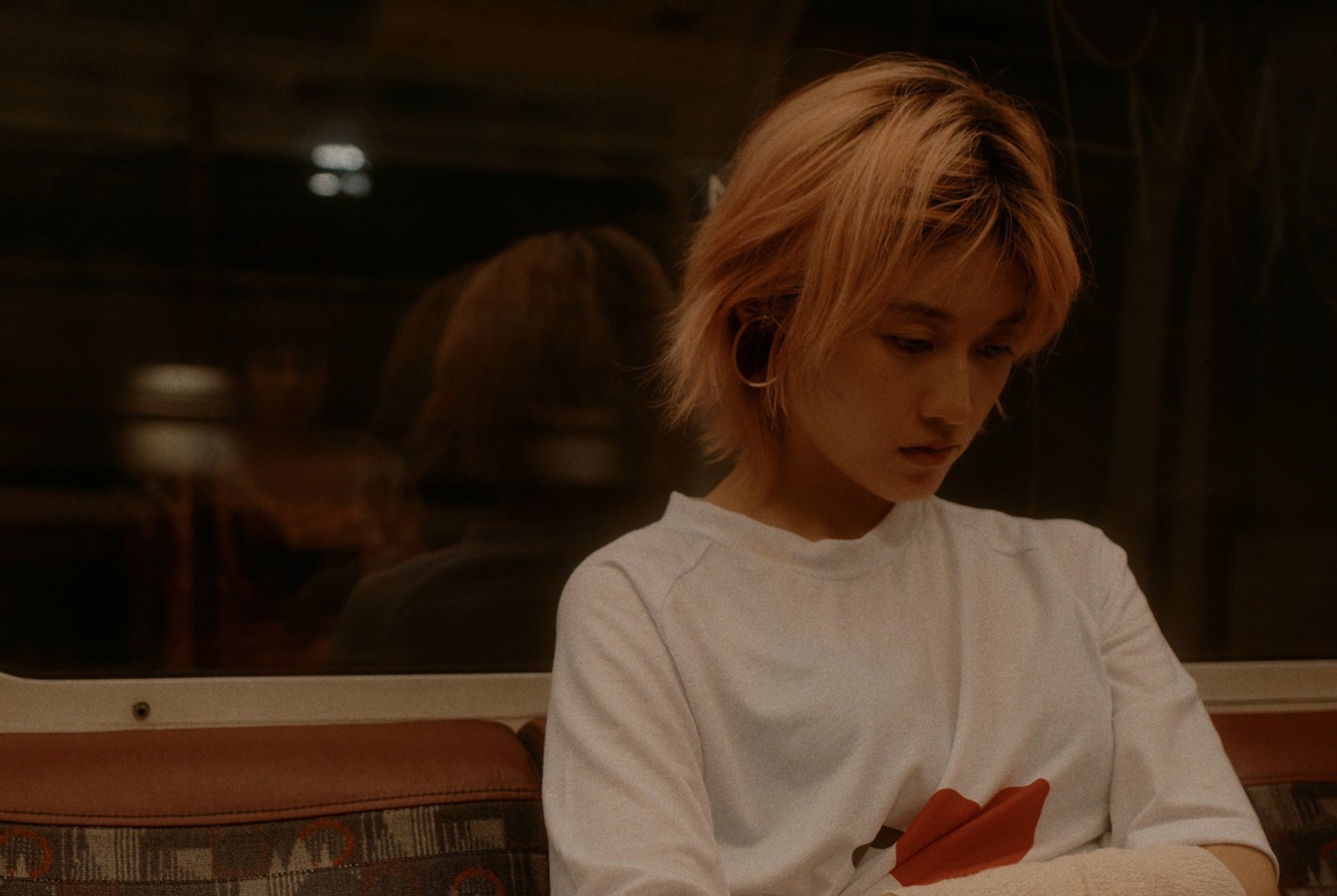 A person with orange hair and a white t-shirt sitting on the train.