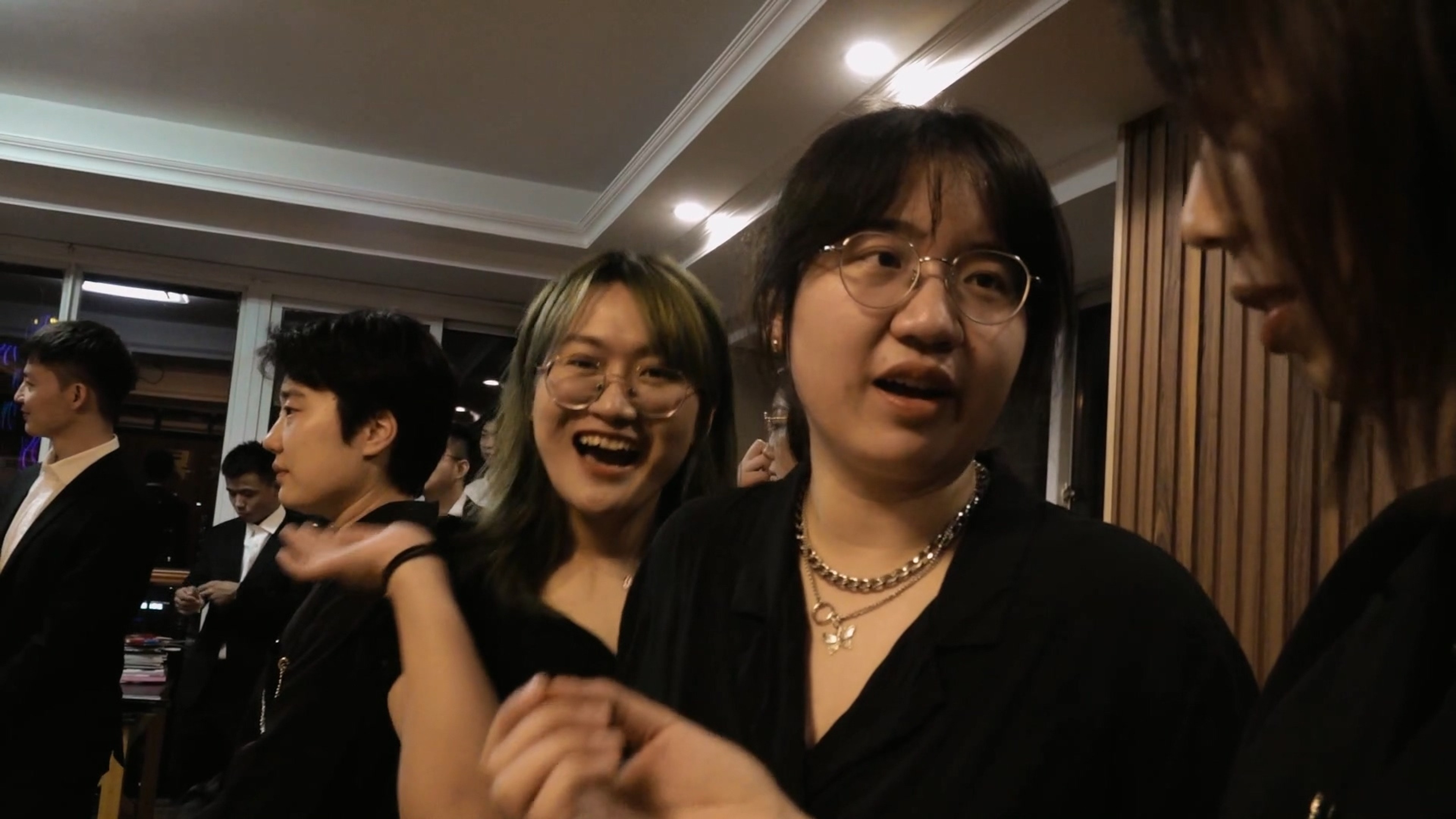 A group of people dressed in black socialising at an indoor gathering, with two smiling figures in the foreground.