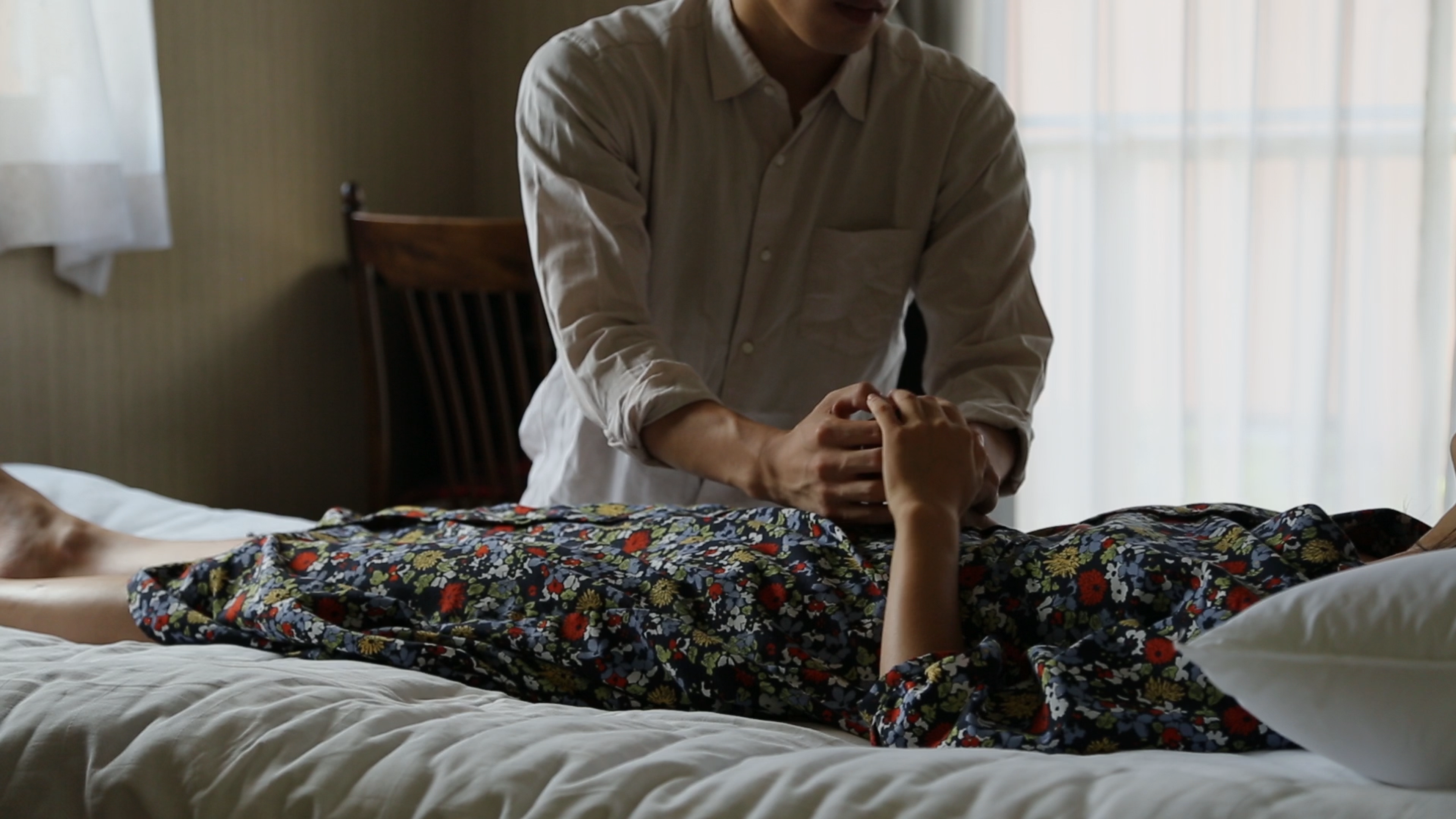 A person lying on a bed in a floral dress while another sits beside them, holding their hand.